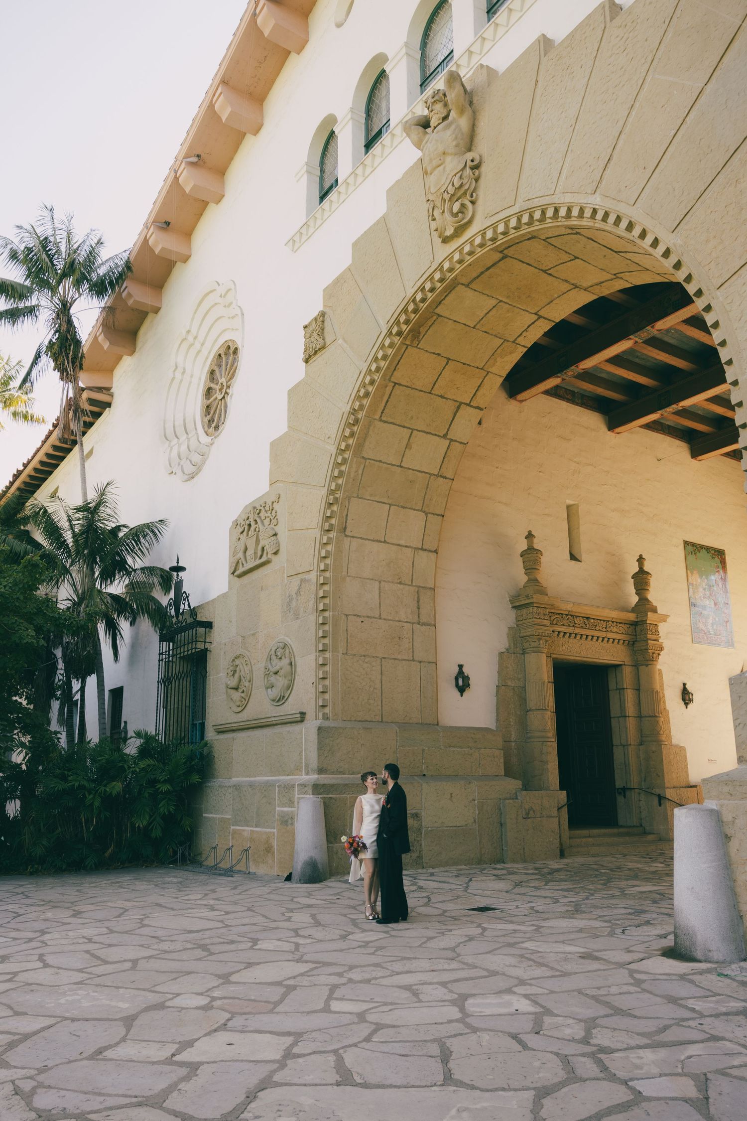 Couple stands beneath ornate stone arch at Santa Barbara Courthouse on their wedding day.