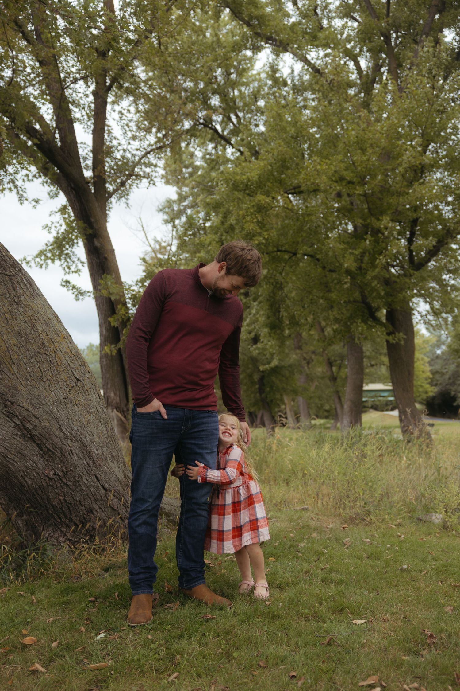 Two people stand together near large tree trunks in a grassy outdoor setting during autumn.