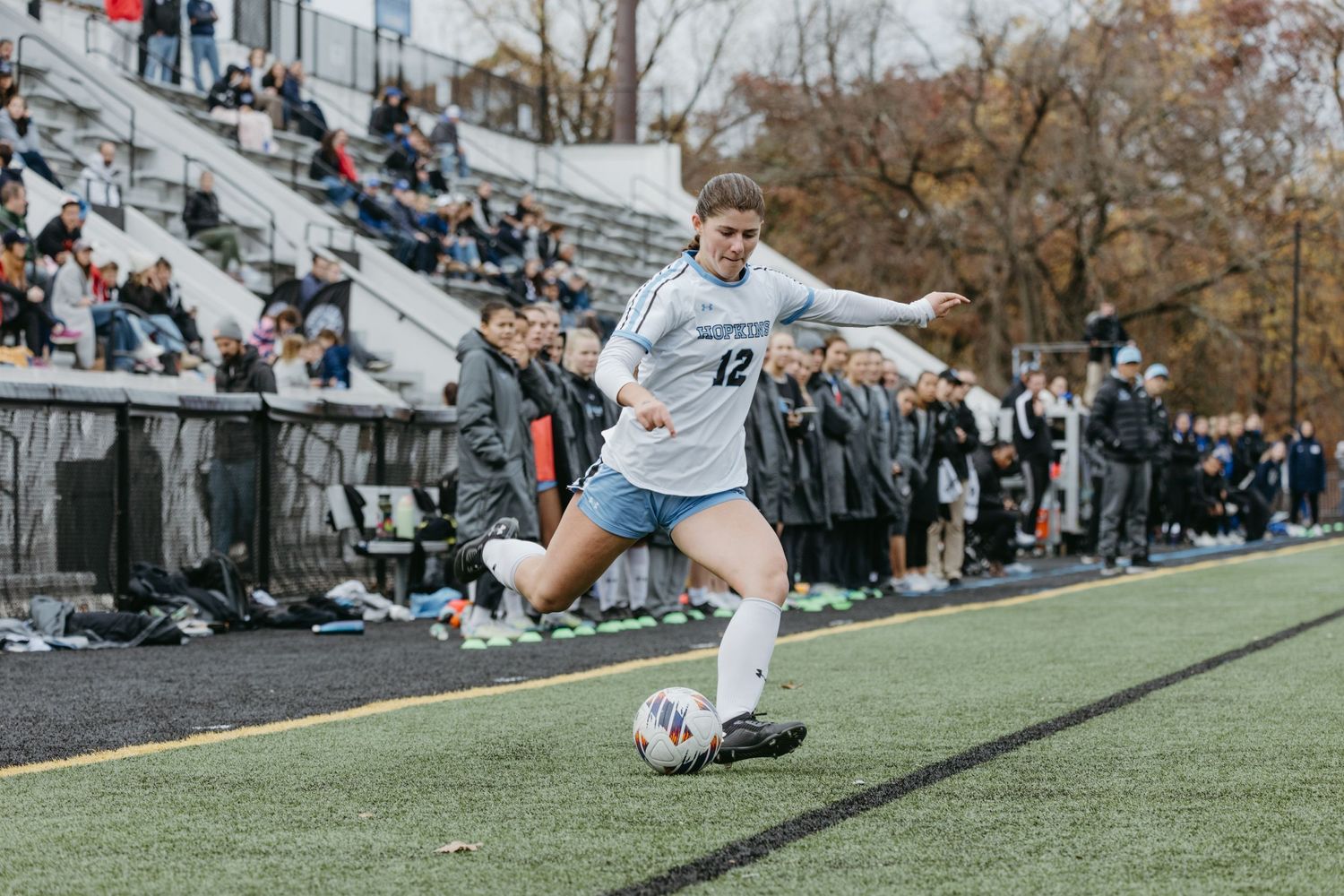 Soccer player maintains ball control while running along sideline with spectators watching.