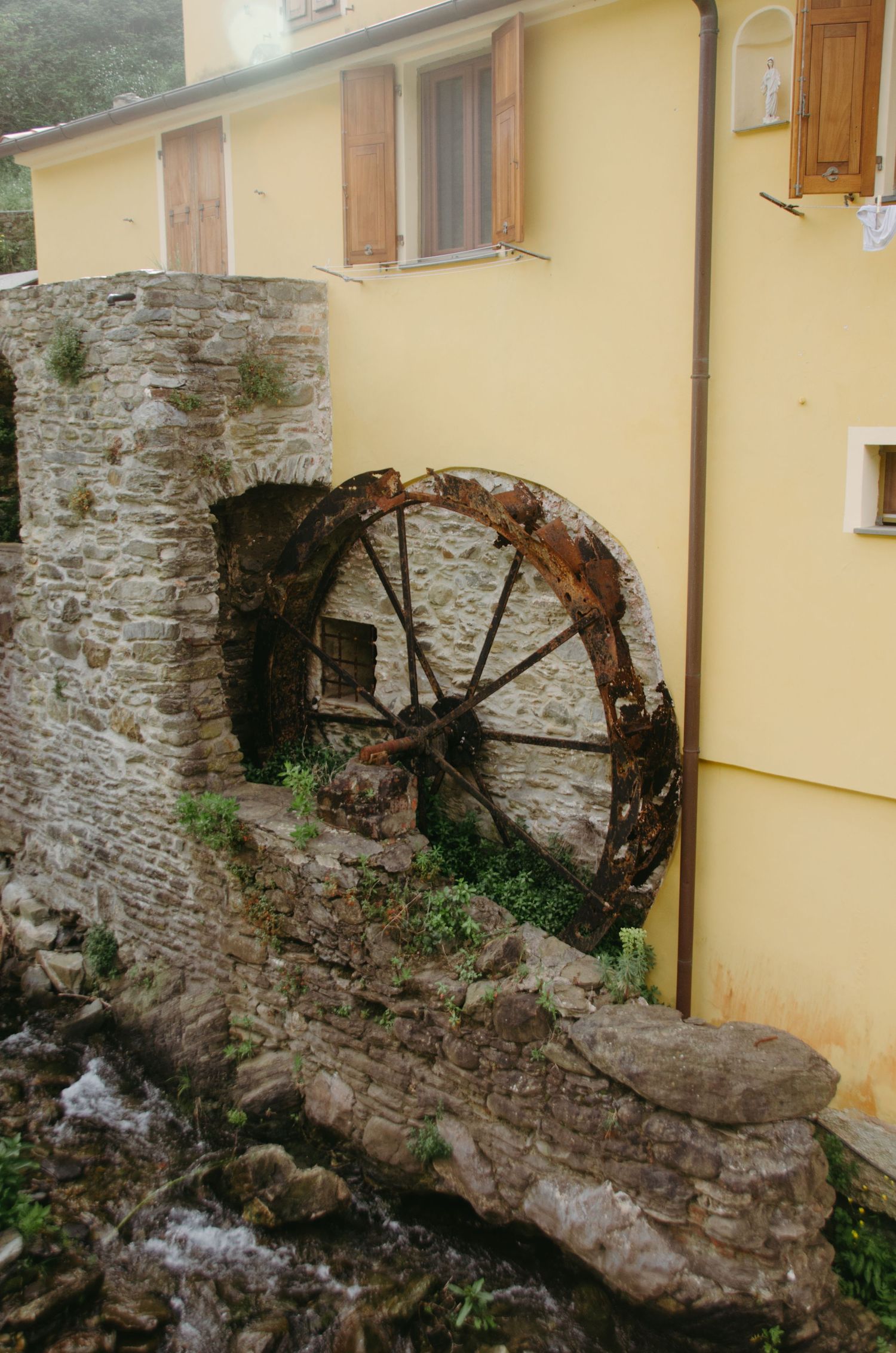 Rustic stone wall with ancient water wheel remains against yellow building exterior.