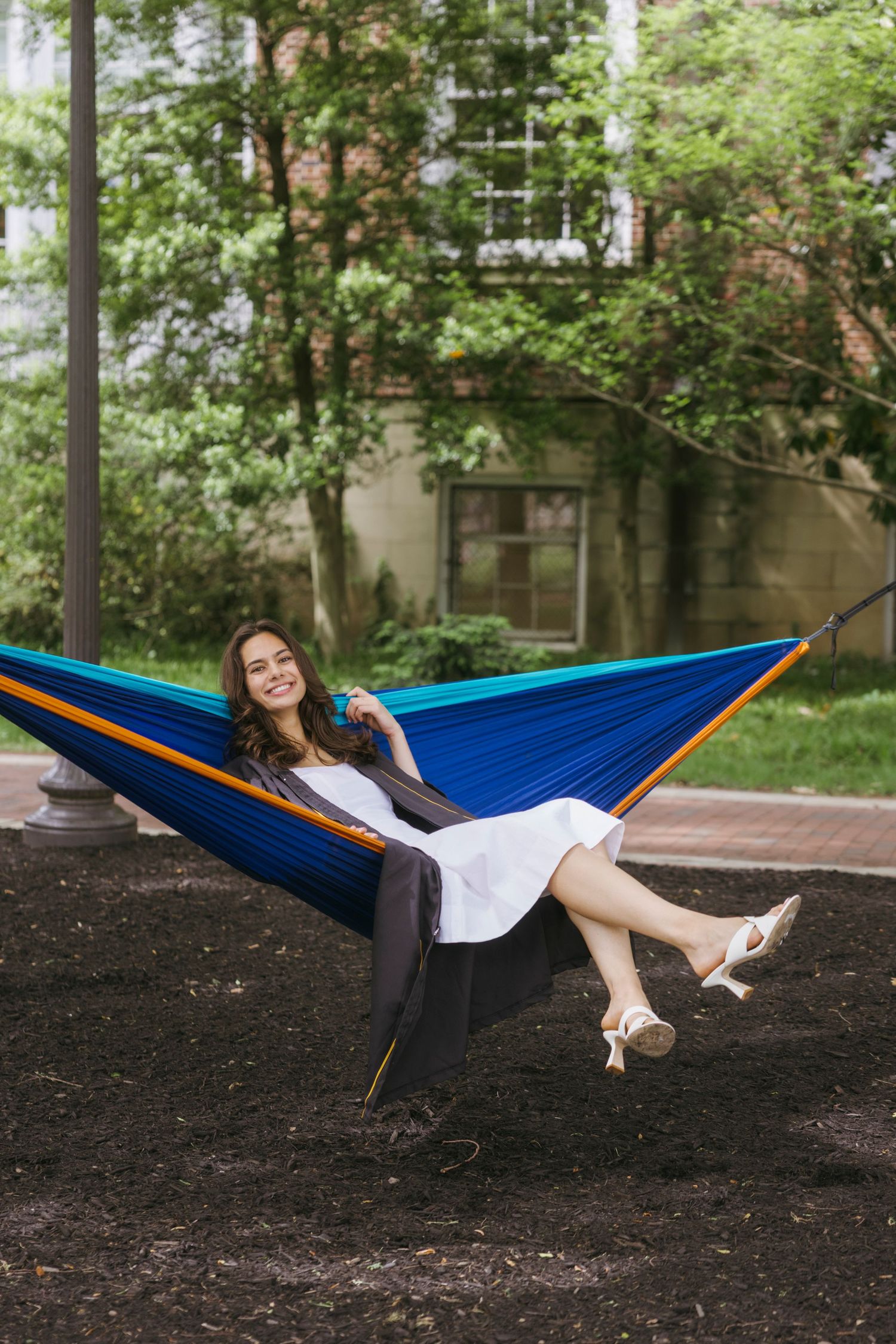 Someone relaxes in a colorful blue and orange camping hammock strung between trees in a peaceful garden setting.