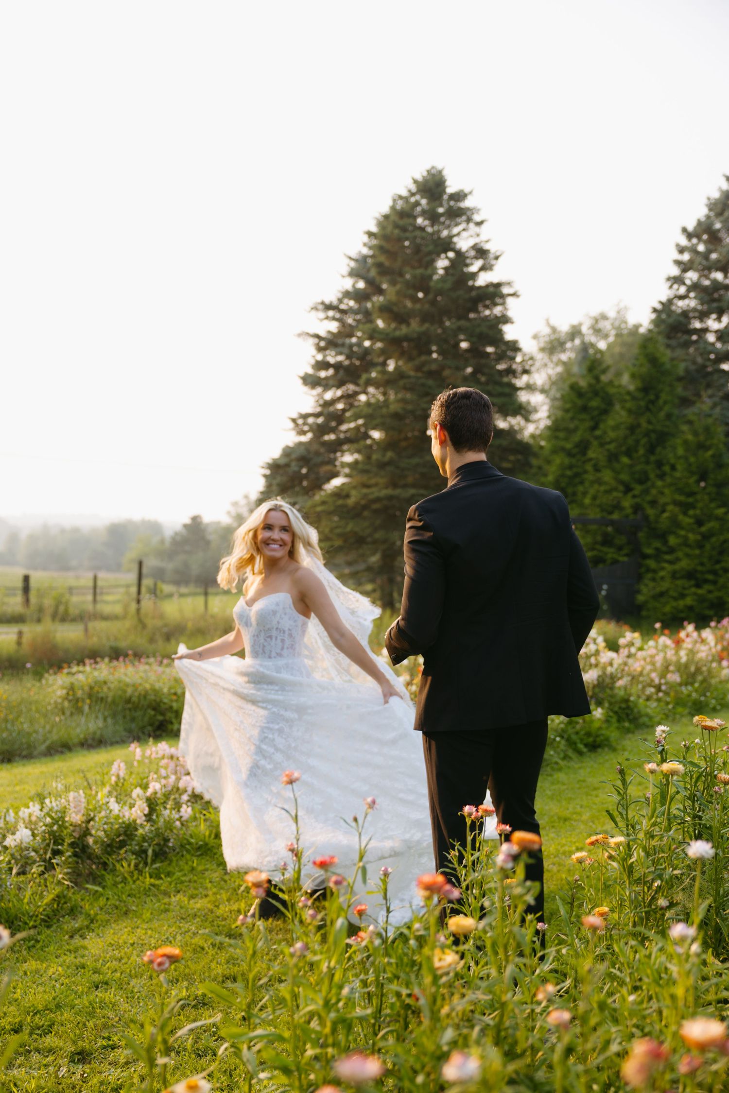 A couple walks through a field of wildflowers and tall grass at sunset while a white dress flows in the breeze.