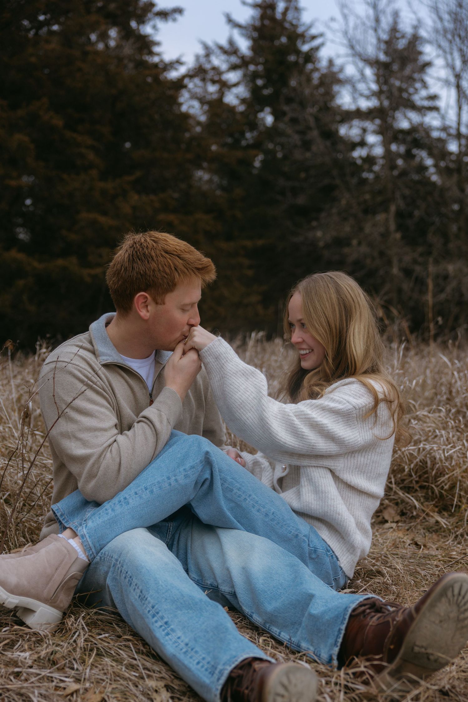 A couple in light sweaters and jeans lying in tall grass during an intimate outdoor photoshoot.
