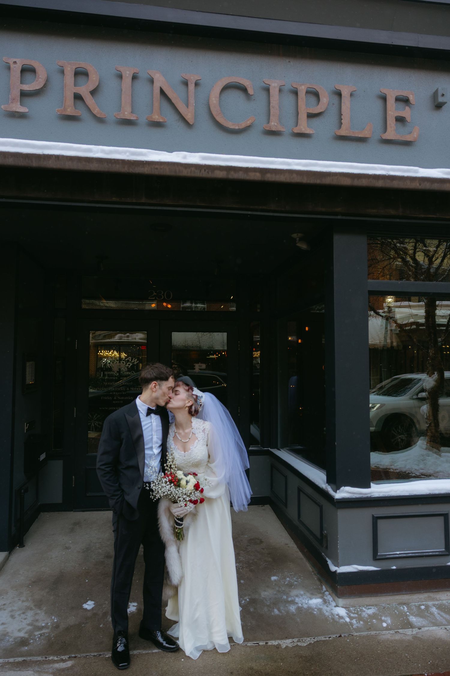 A couple in wedding attire share a romantic moment outside a storefront with 'PRINCIPLE' signage.