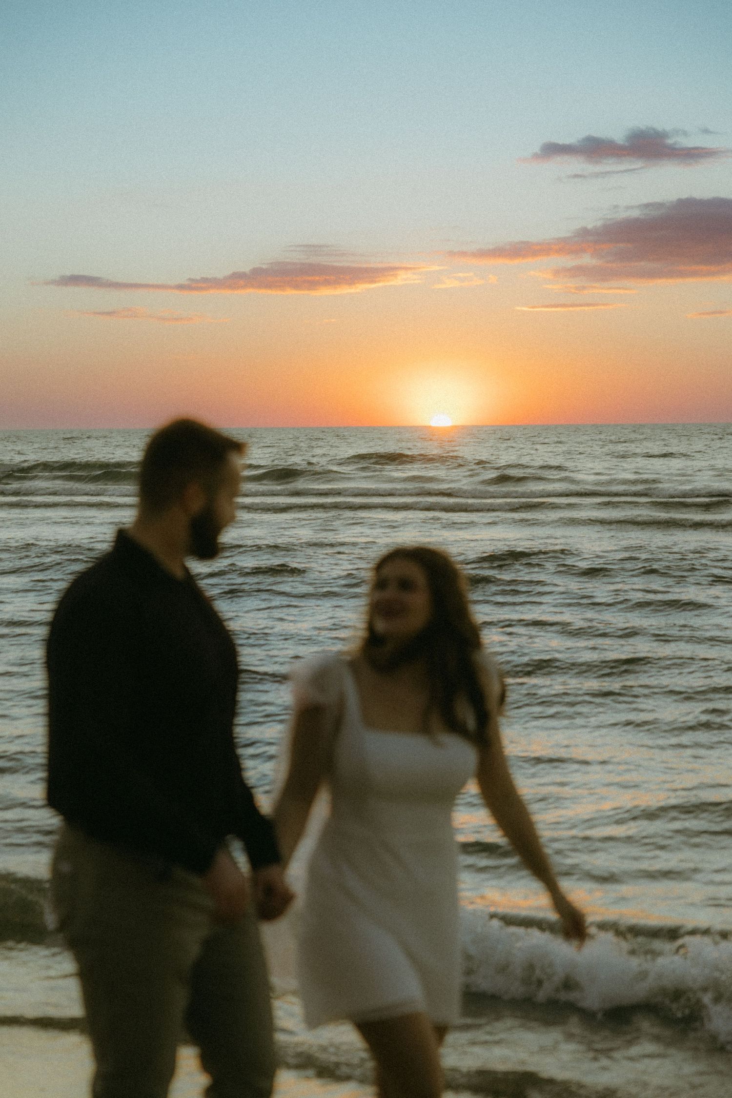 A couple dances together on a beach at sunset with waves crashing behind them in a romantic seaside moment.