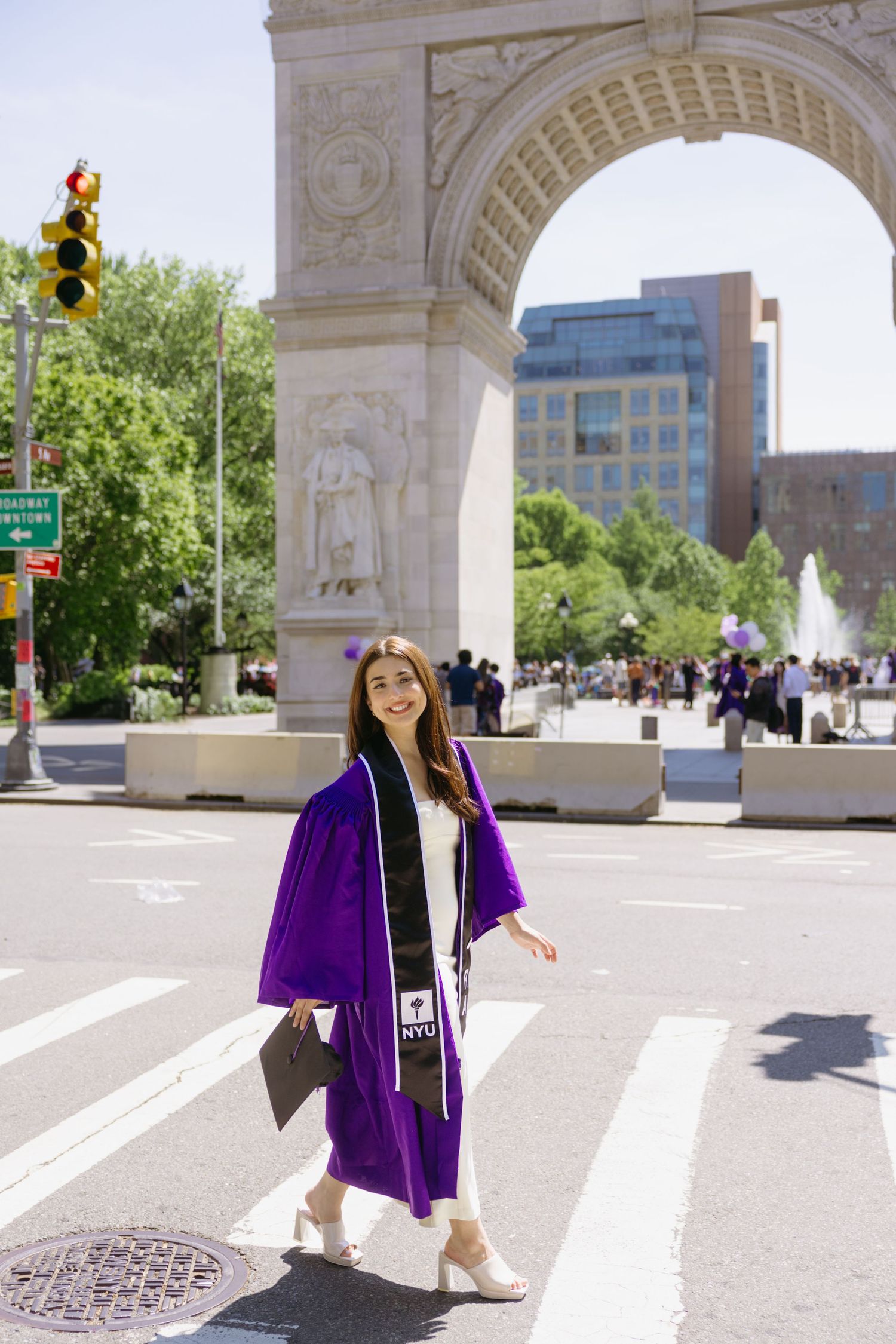 A person in graduation attire poses in front of Washington Square Arch in New York City on a sunny day.