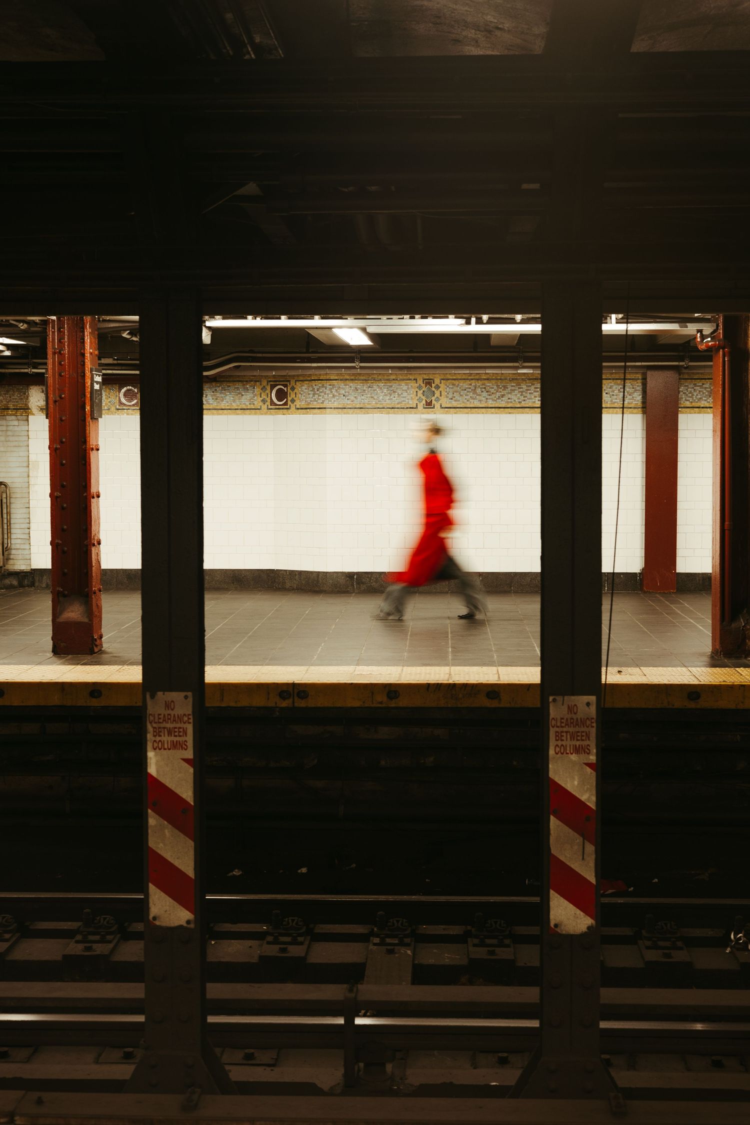 A blurred figure in red walks past white tiled walls in a New York City subway station platform between dark pillars.