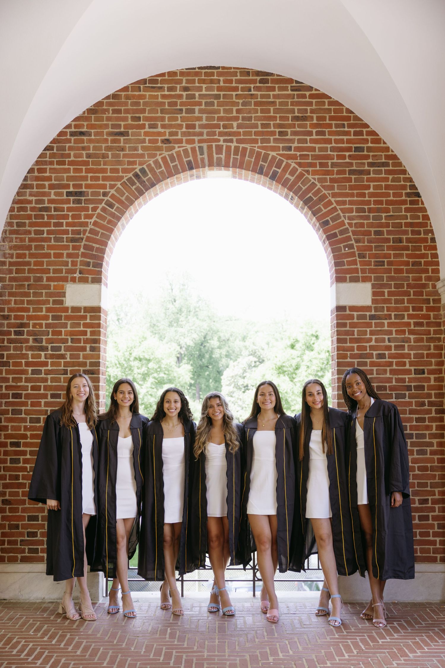 Group in black graduation gowns and white dresses standing under a brick archway on campus.