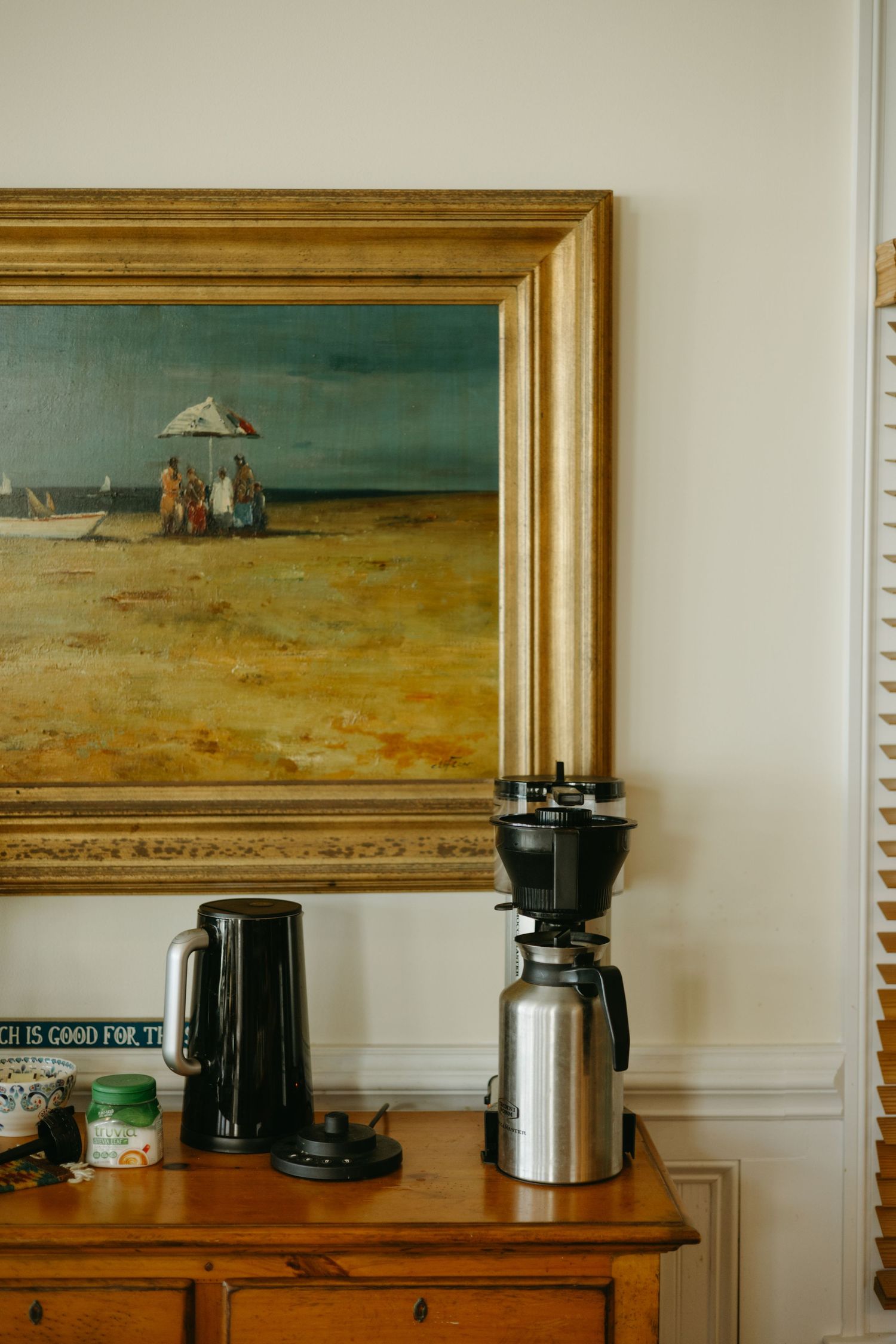 A coffee making station with electric kettle and grinder on wooden cabinet beneath framed beach scene artwork.