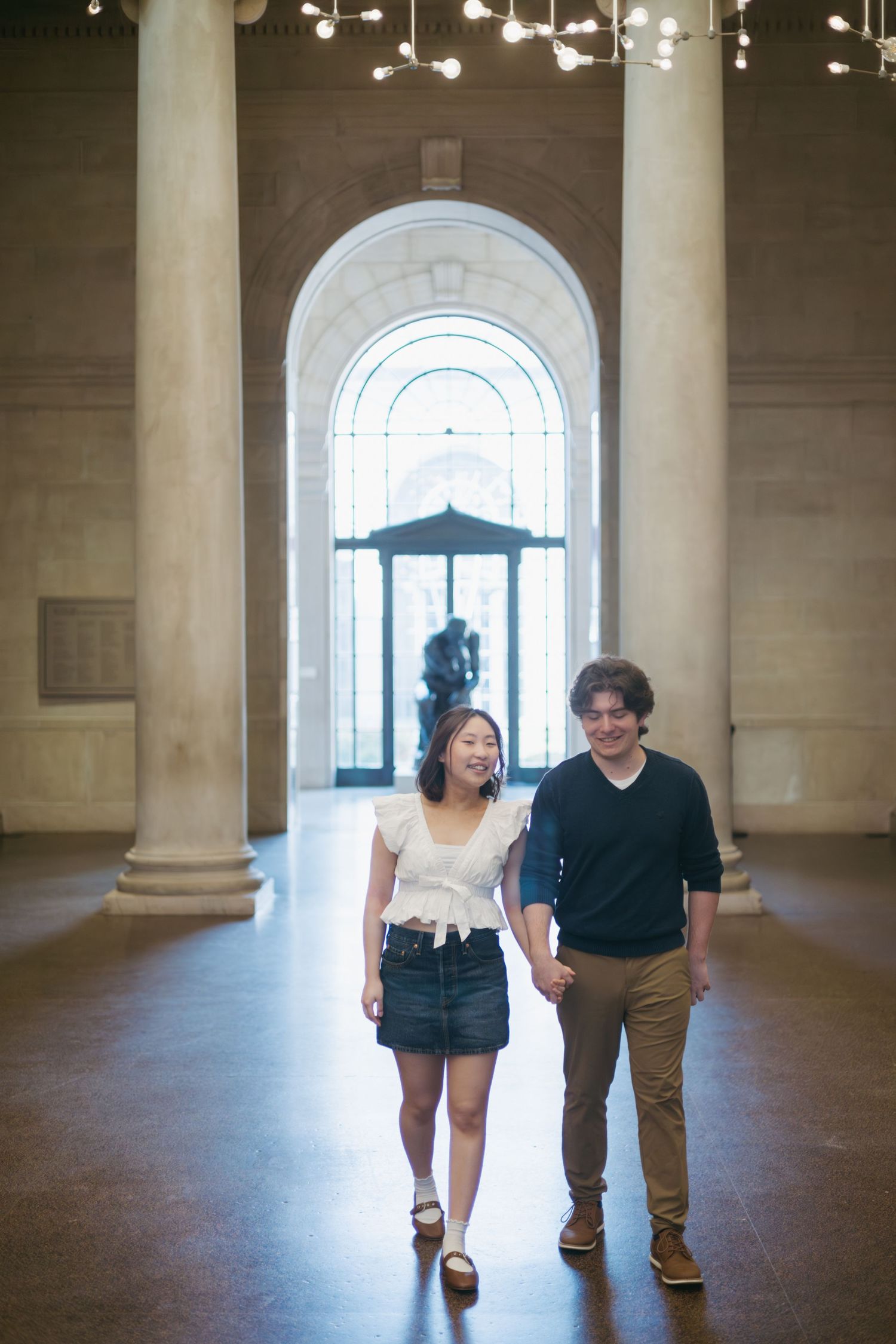 A couple walks together through an elegant building hallway with arched windows and columns, bathed in natural light.