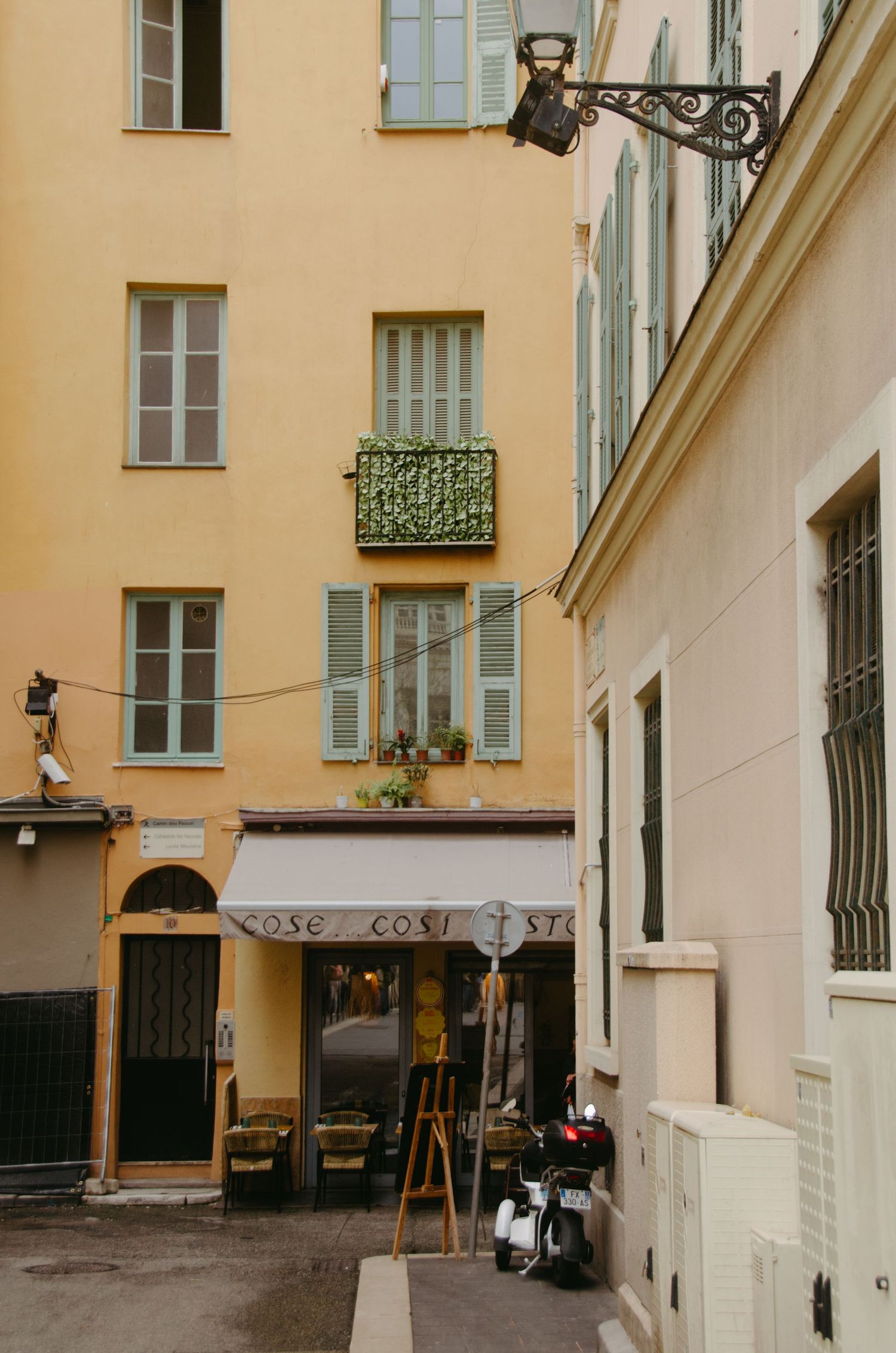 Narrow street view of pastel colored buildings with blue shutters and ornate iron balconies in a European city.