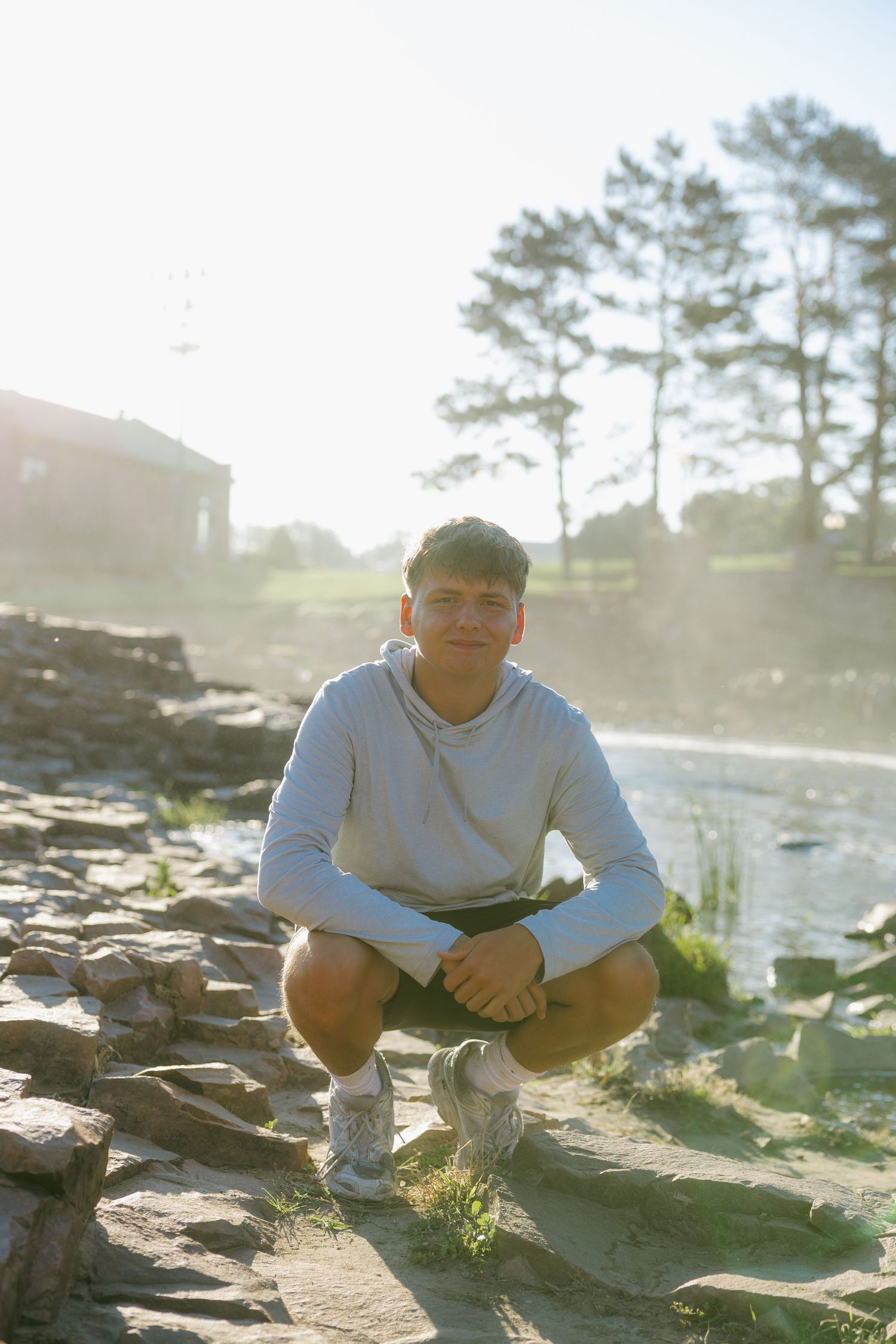 Young man crouching on rocky shore near water, wearing gray hoodie, with trees and building in sunny background.