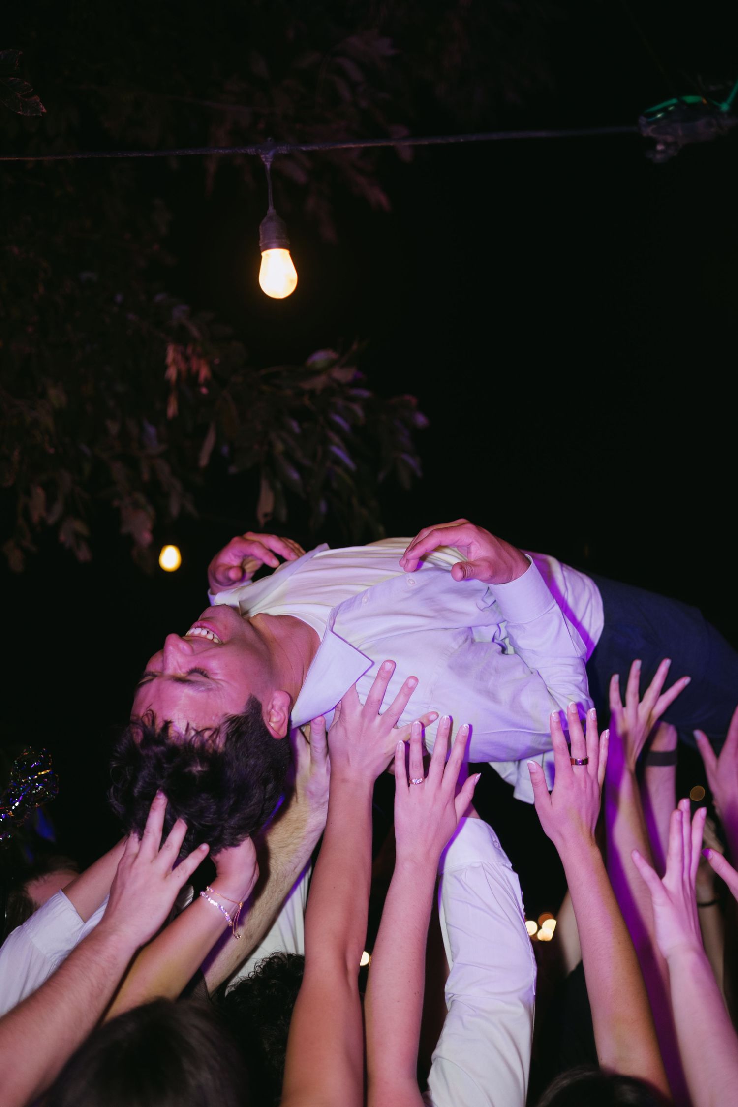 Bearded groom crowd surfing at outdoor wedding reception at night with string lights above.