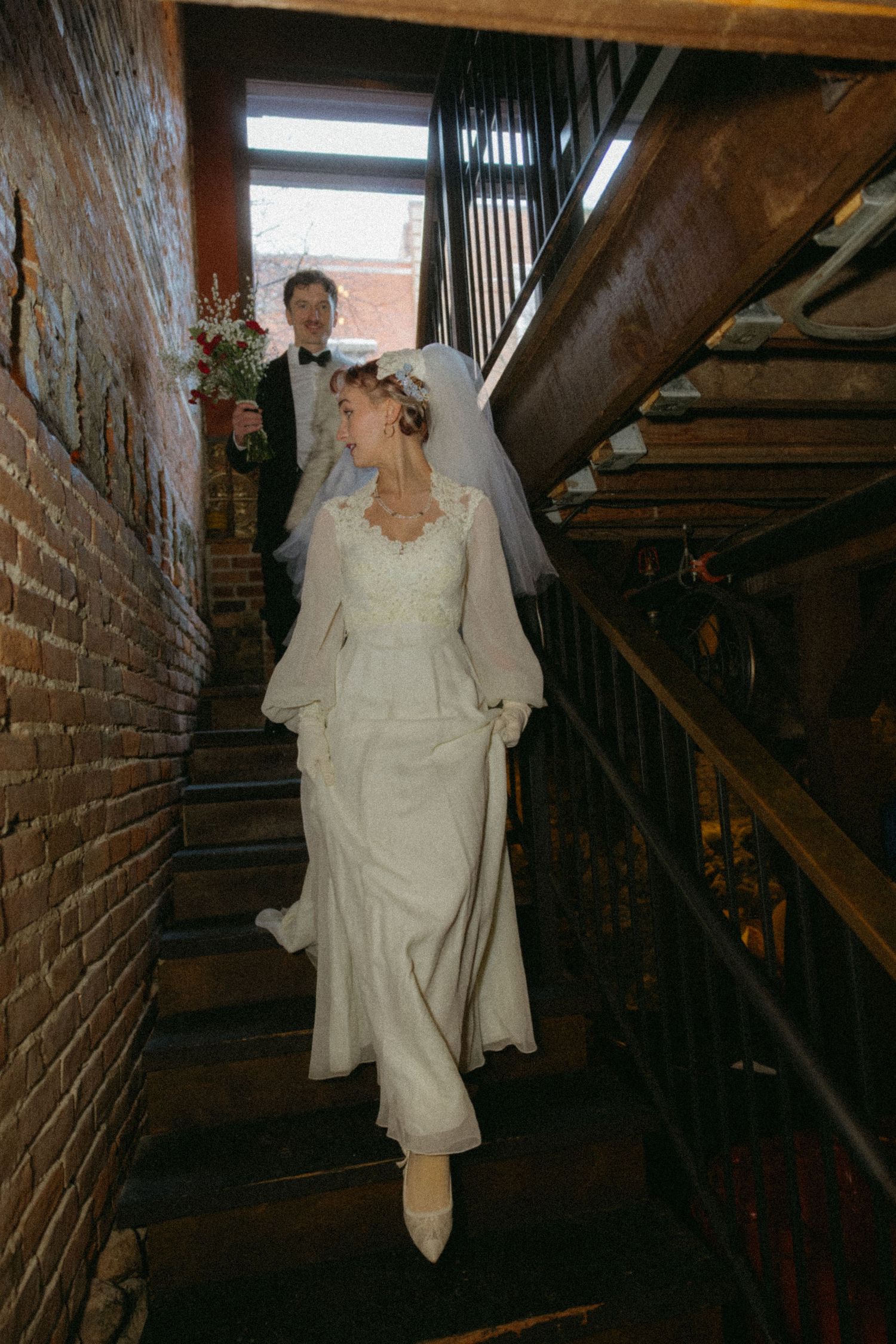 A person in a white wedding dress walks down a dimly lit brick corridor with wooden beams overhead.