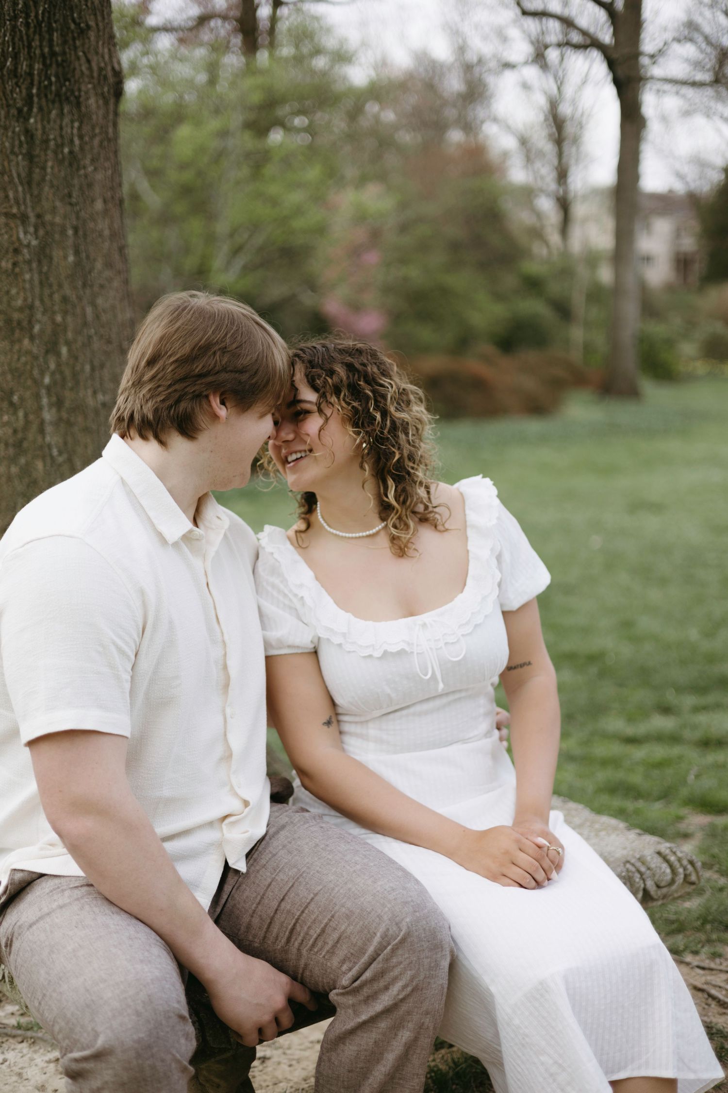 A romantic couple in white outfits sitting close together outdoors in a park setting during springtime.