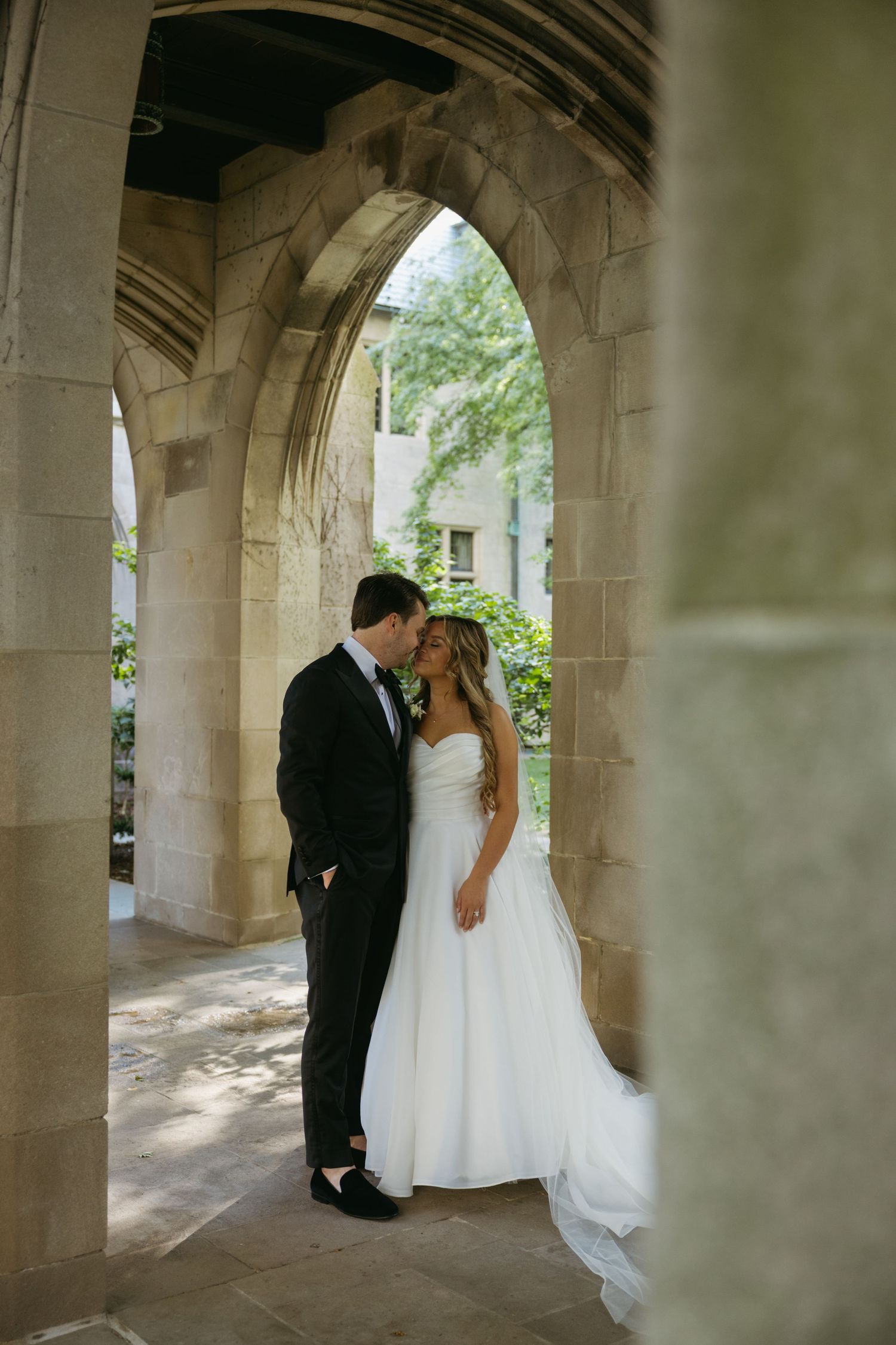 A couple in wedding attire embraces beneath elegant stone arches.