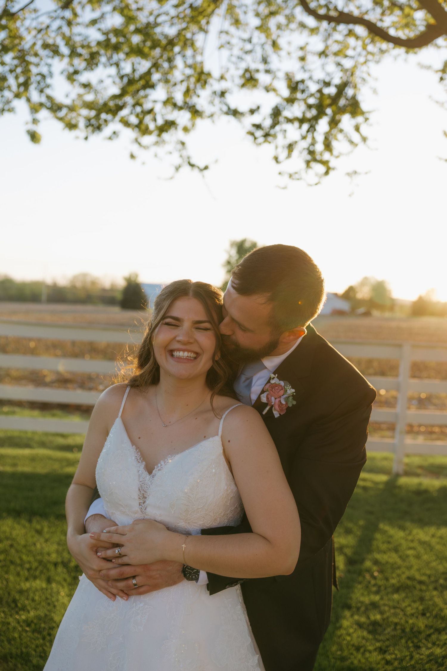 Bride and groom embracing at golden hour sunset outdoors, white fence and green fields in background.