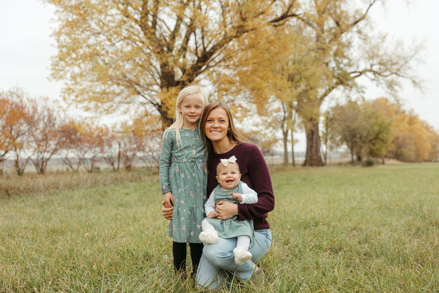 A family in casual attire embraces while standing in a grassy field during autumn with golden trees in the background.