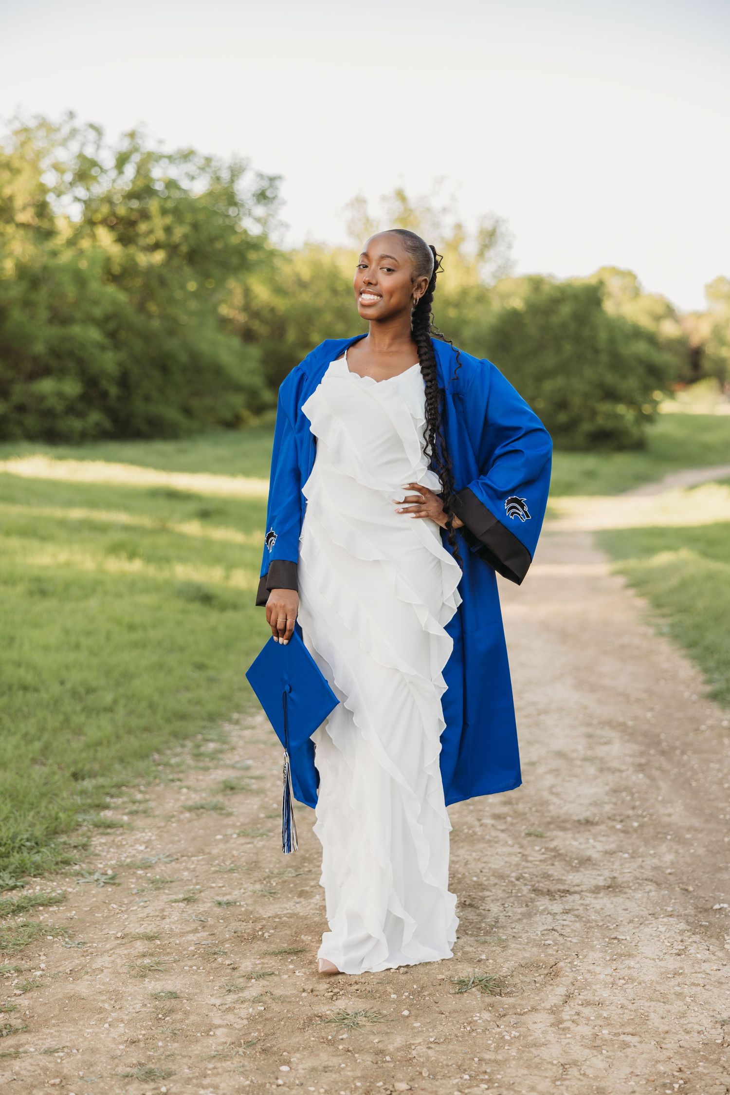 Joyful Black female graduate in blue robe and white dress walking along scenic outdoor path.