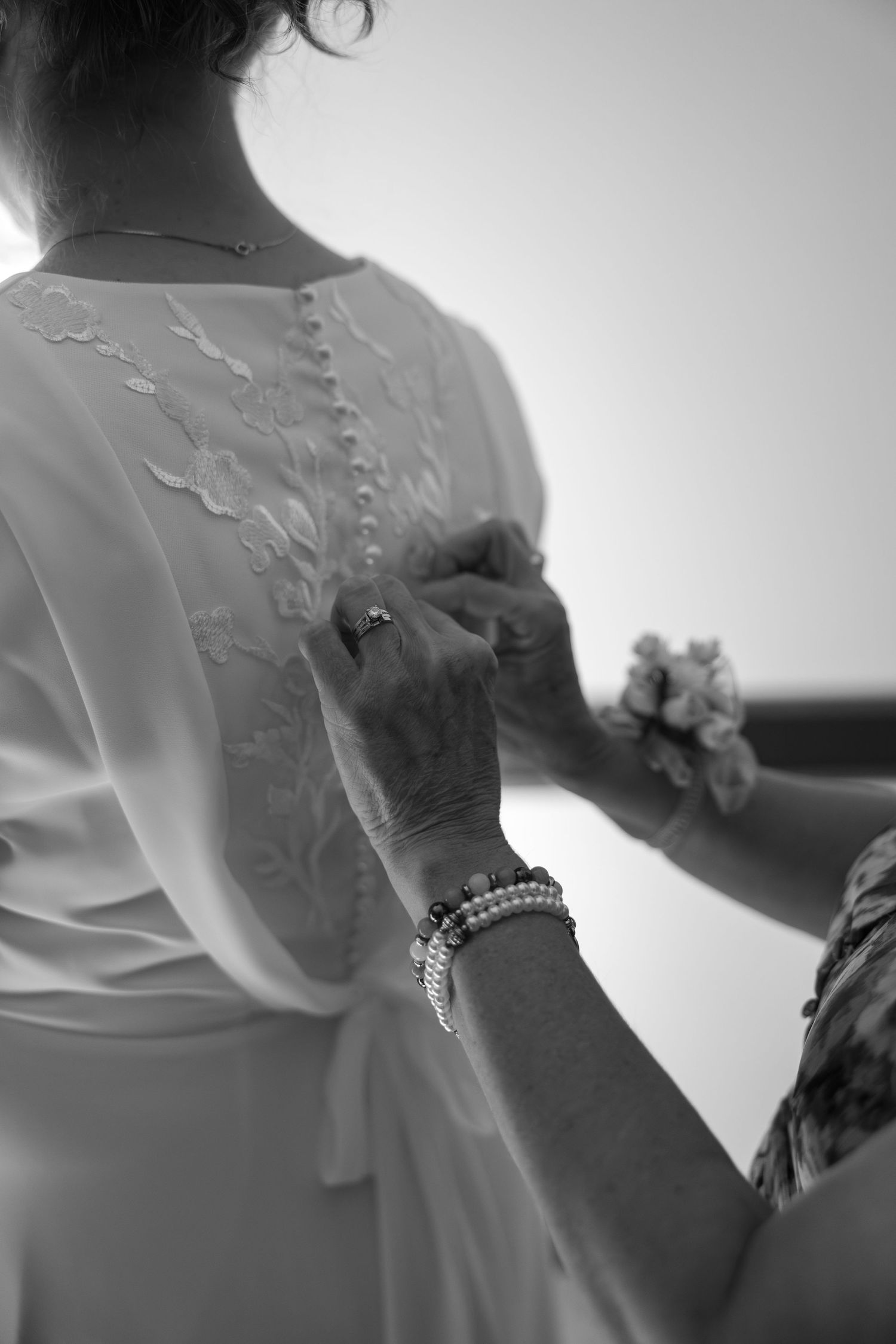 Black and white photo of a woman having the buttons of her lace wedding dress fastened by a helper.