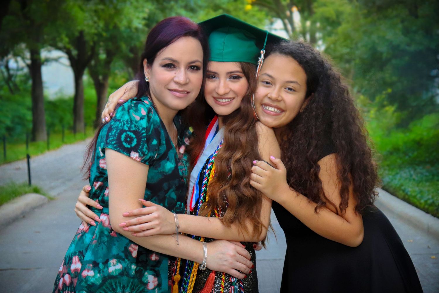 Three young women embrace and smile together outdoors during graduation celebration in green cap and gown.