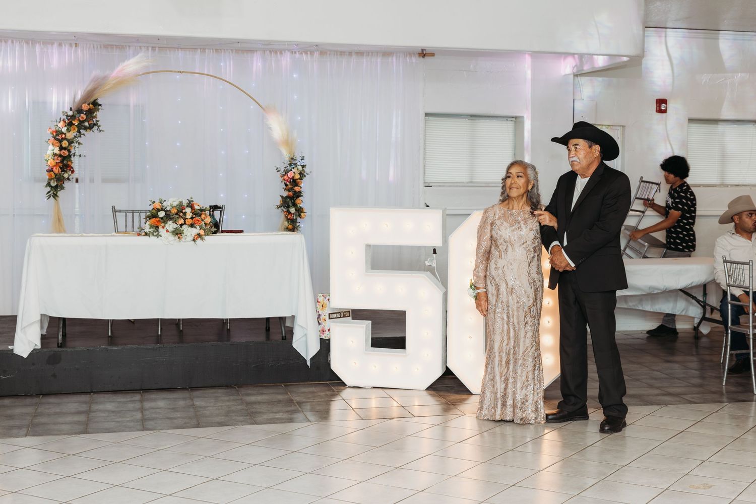 Couple poses beside large number 50 display at golden anniversary celebration in decorated hall.
