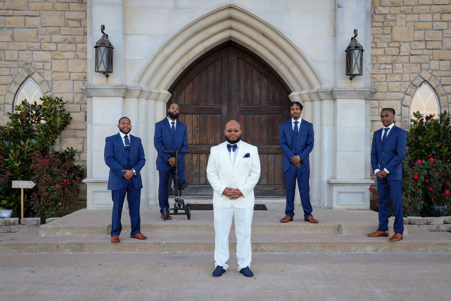 Groom in white suit with groomsmen in blue suits outside stone church.