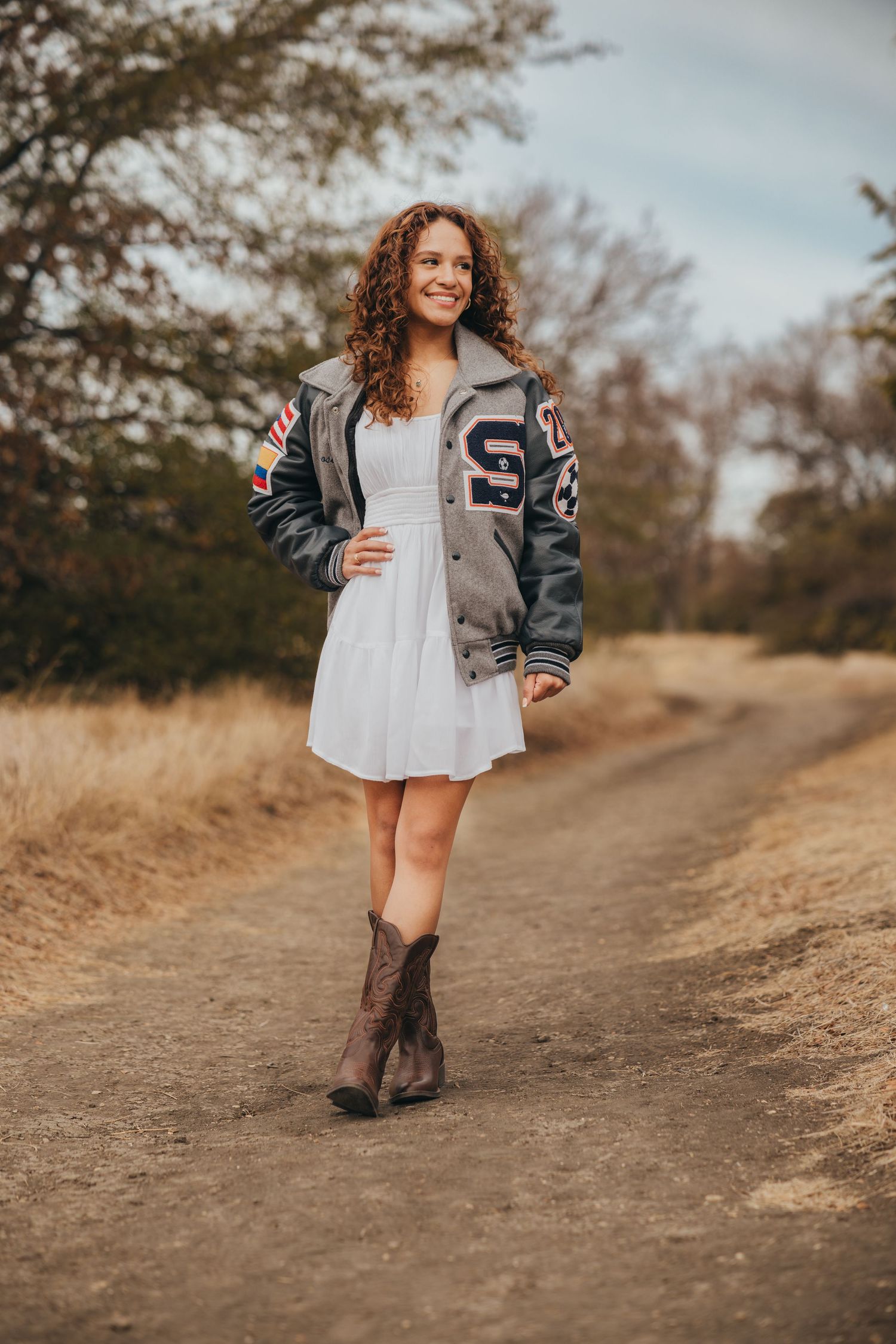 Young woman in varsity jacket white dress and cowboy boots on dirt road senior portrait.