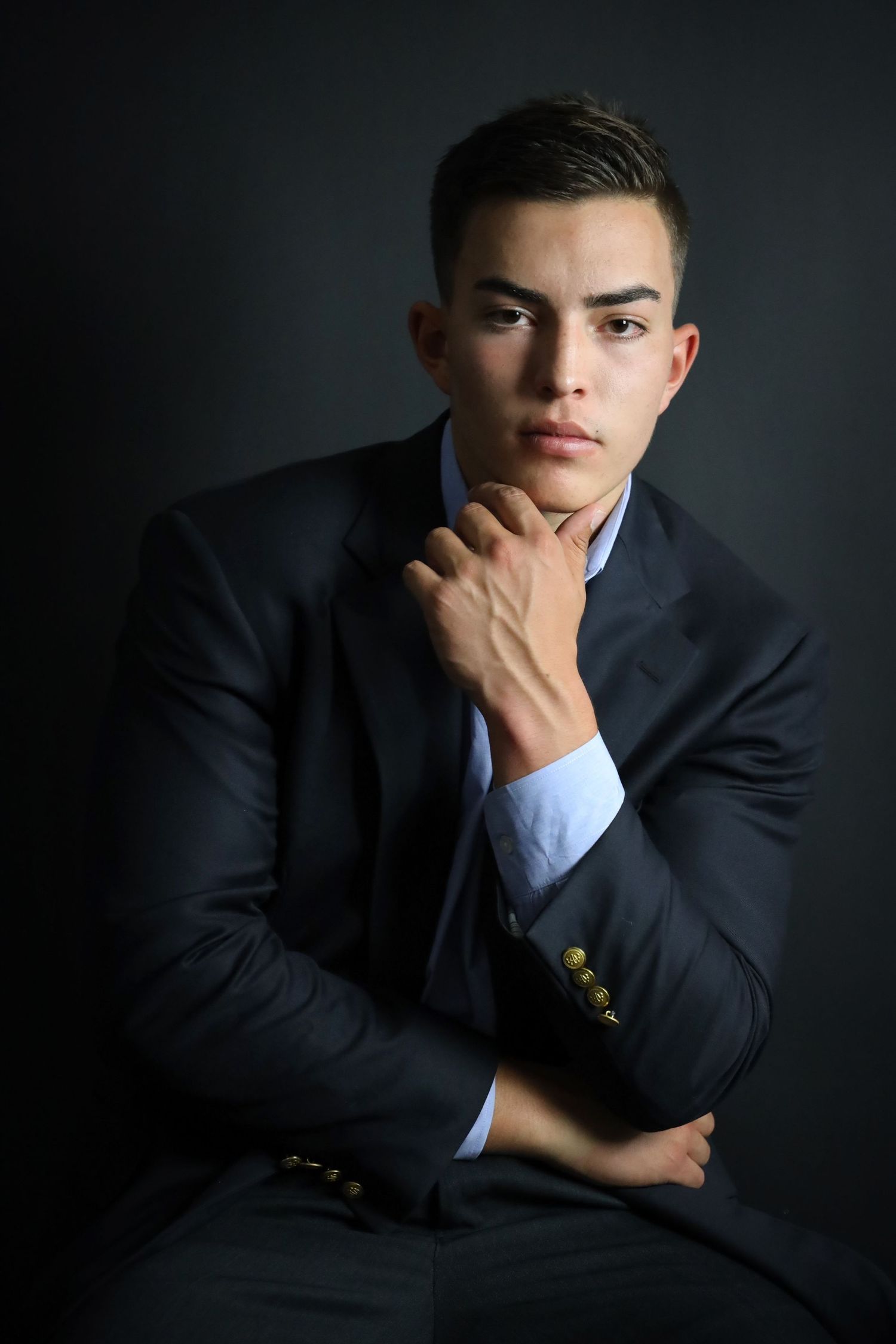 Young man in dark suit posing thoughtfully against black background.