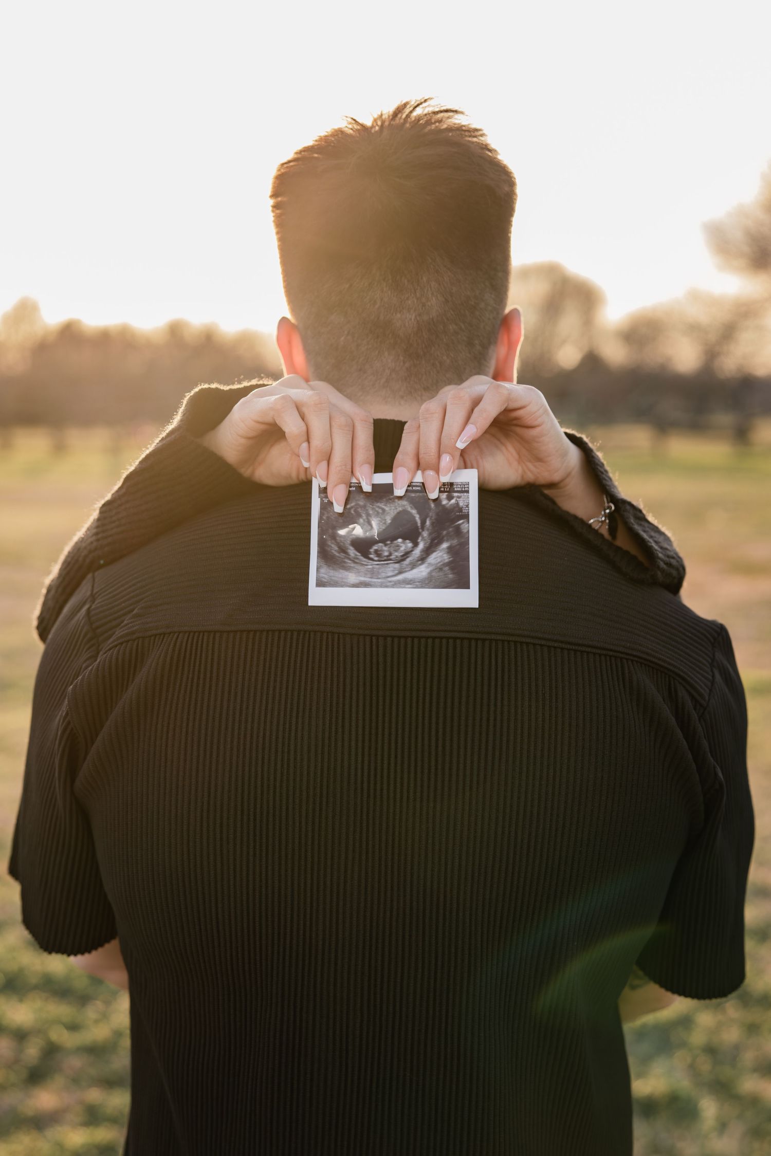Man holding an ultrasound photo behind his back in a sunny outdoor field.