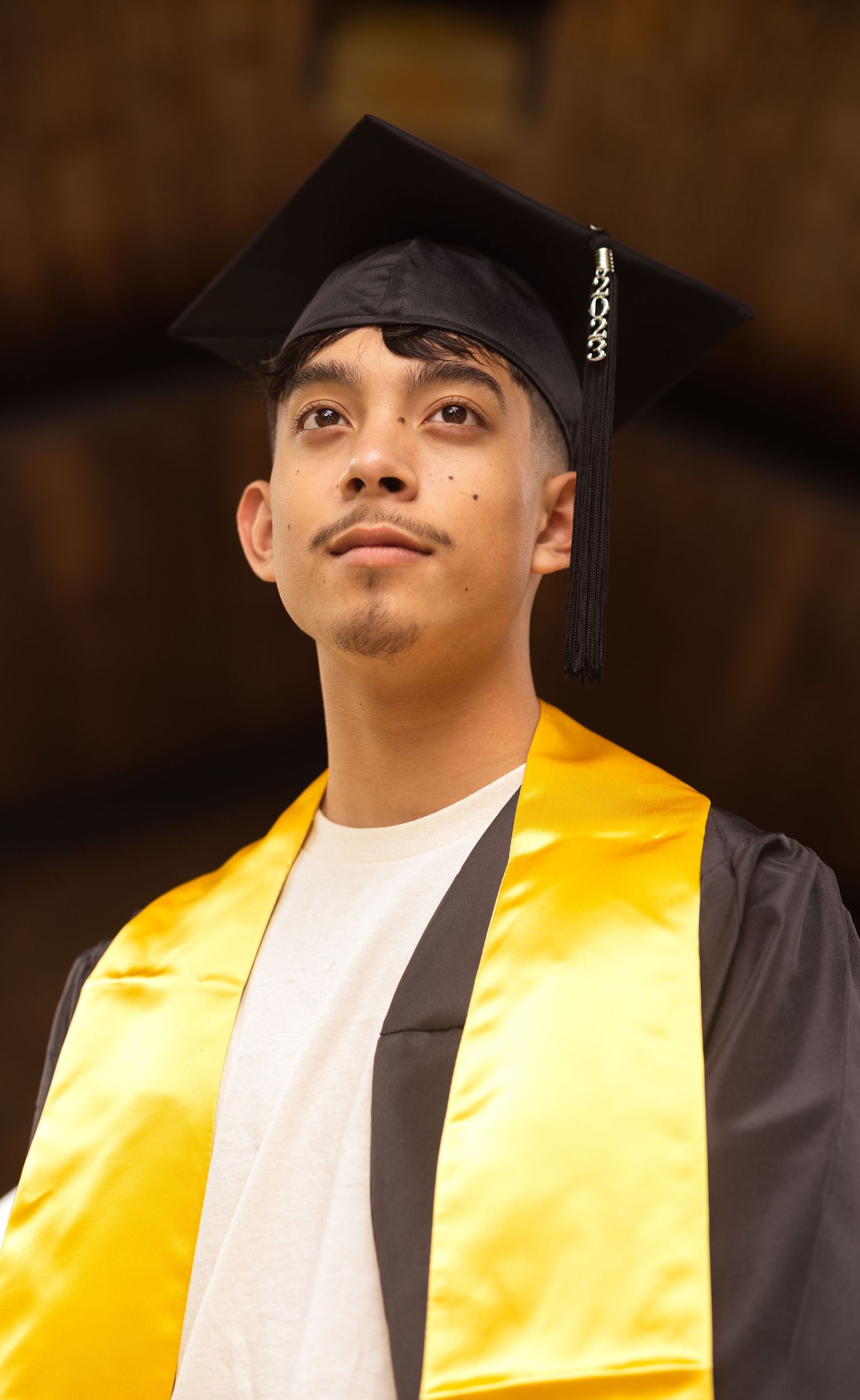 Young male graduate in black cap and gold stole looking upward in dramatic lighting.