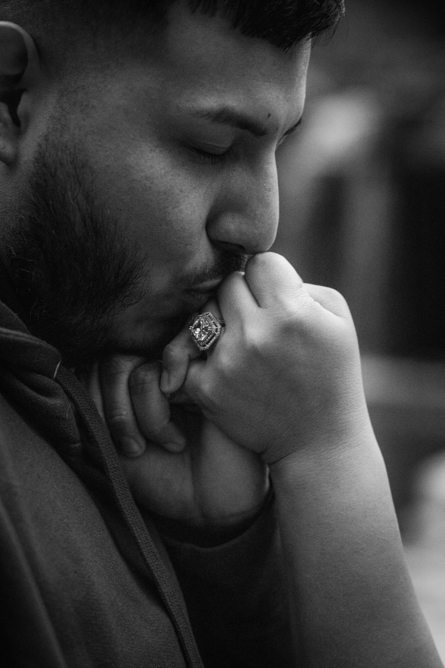 Black and white portrait of bearded man kissing ornate ring held in clasped hands with eyes closed.