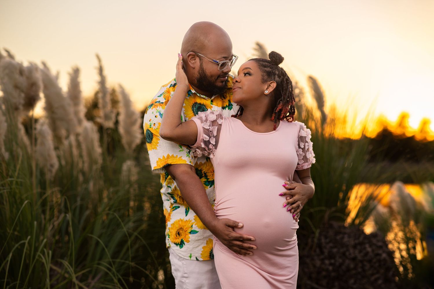 Expectant couple shares tender forehead kiss at sunset in field of tall pampas grass, man in floral shirt.