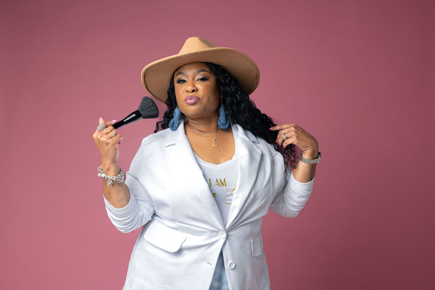 Confident plus-size woman in white blazer and tan hat holding makeup brushes on pink background.