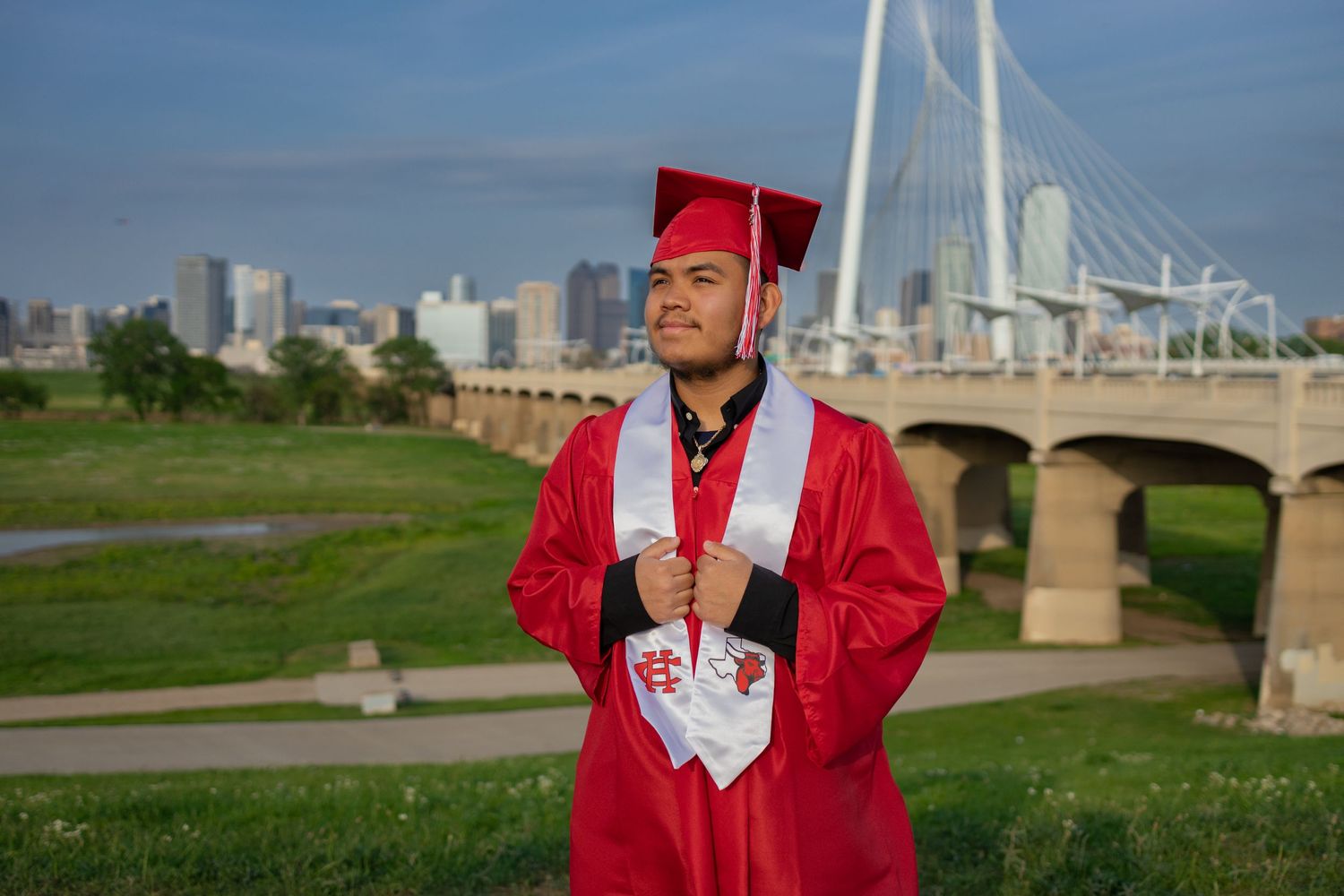 Male graduate in red cap and gown poses with city bridge skyline in background outdoors.