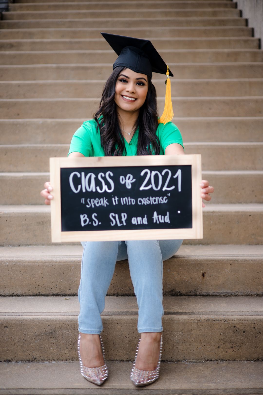 Graduate sits on steps holding Class of 2021 B.S. SLP and Aud chalkboard sign wearing black cap.