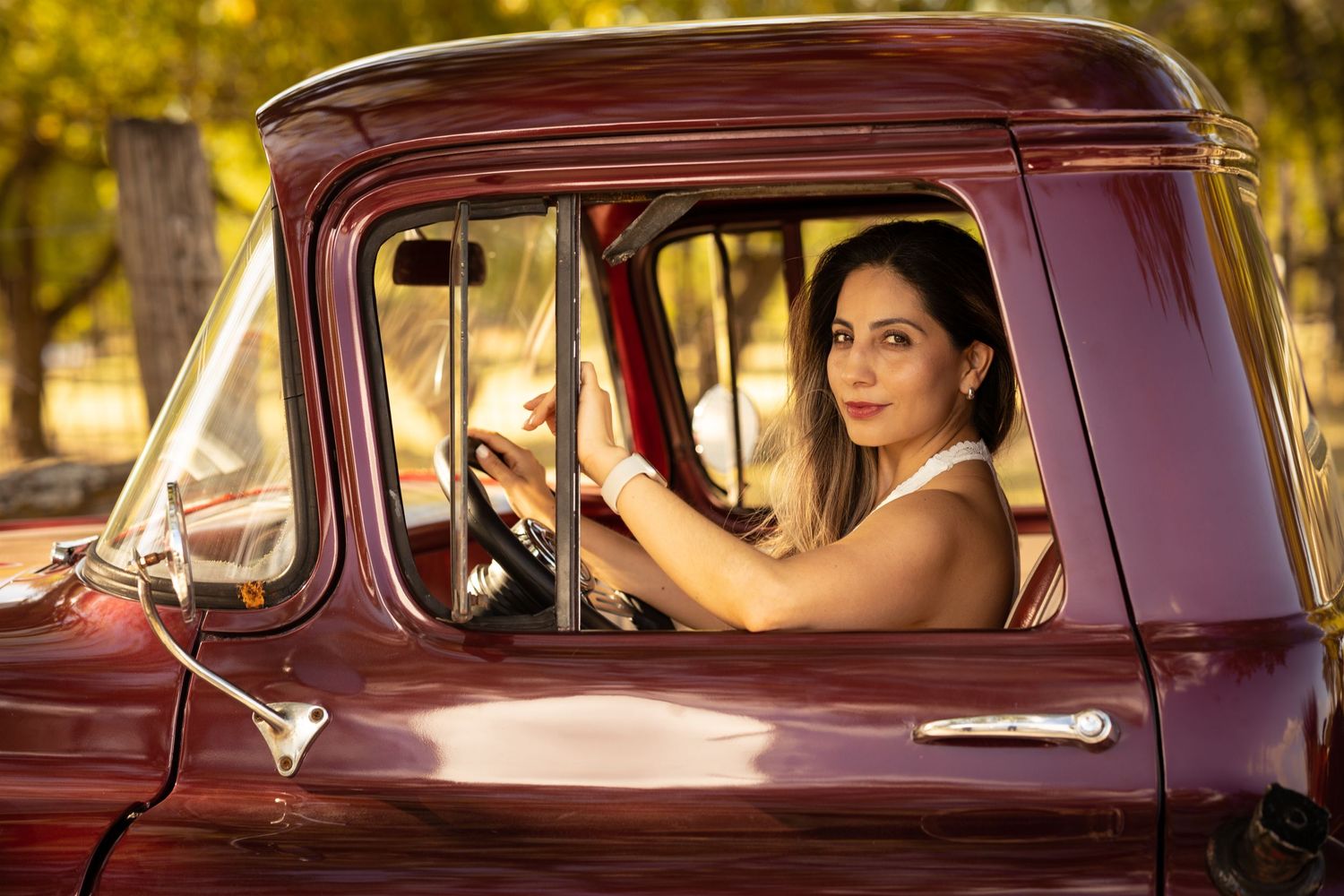 Smiling woman in white sleeveless dress sits behind wheel of classic burgundy pickup truck in sunny outdoor setting.