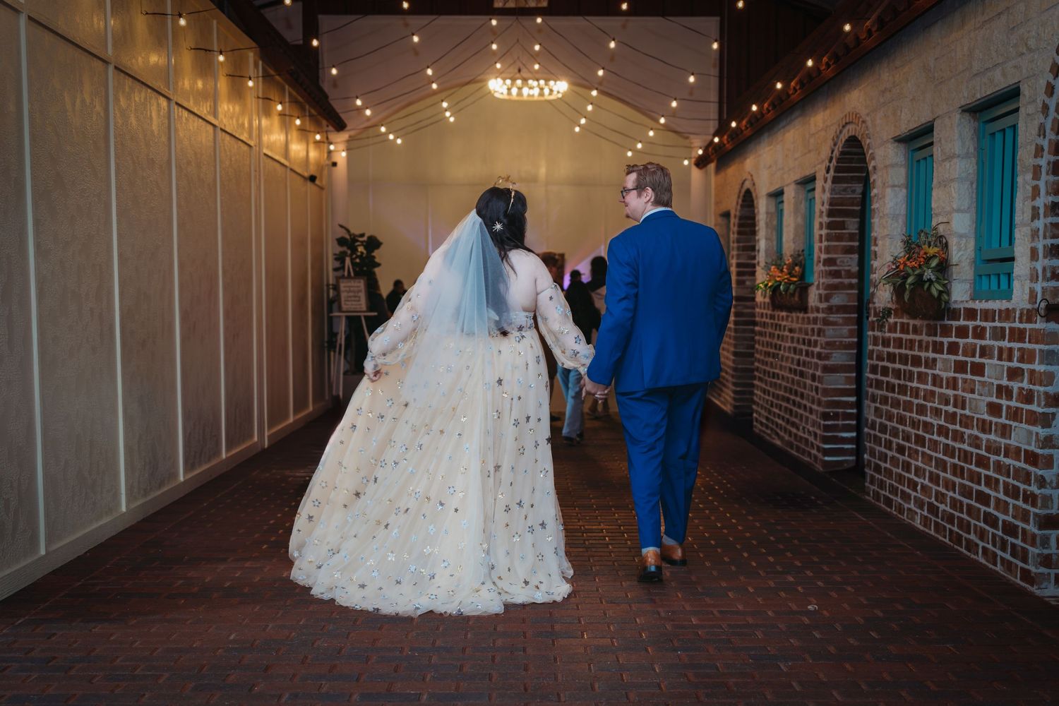 Bride in a floral wedding gown and groom in blue suit walking hand in hand through a string-lit corridor.