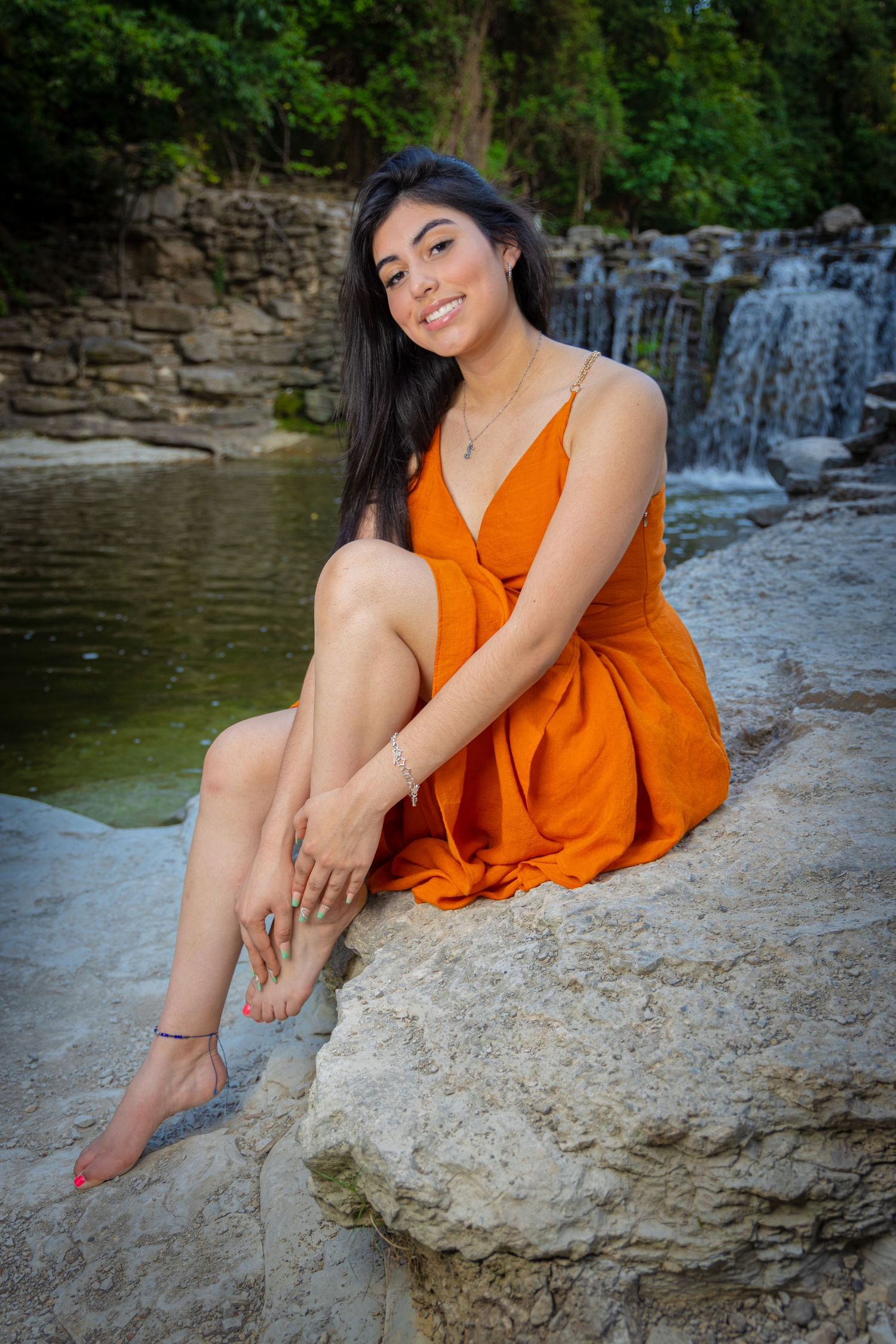 Smiling young woman in orange sundress sitting on rocks near scenic waterfall.