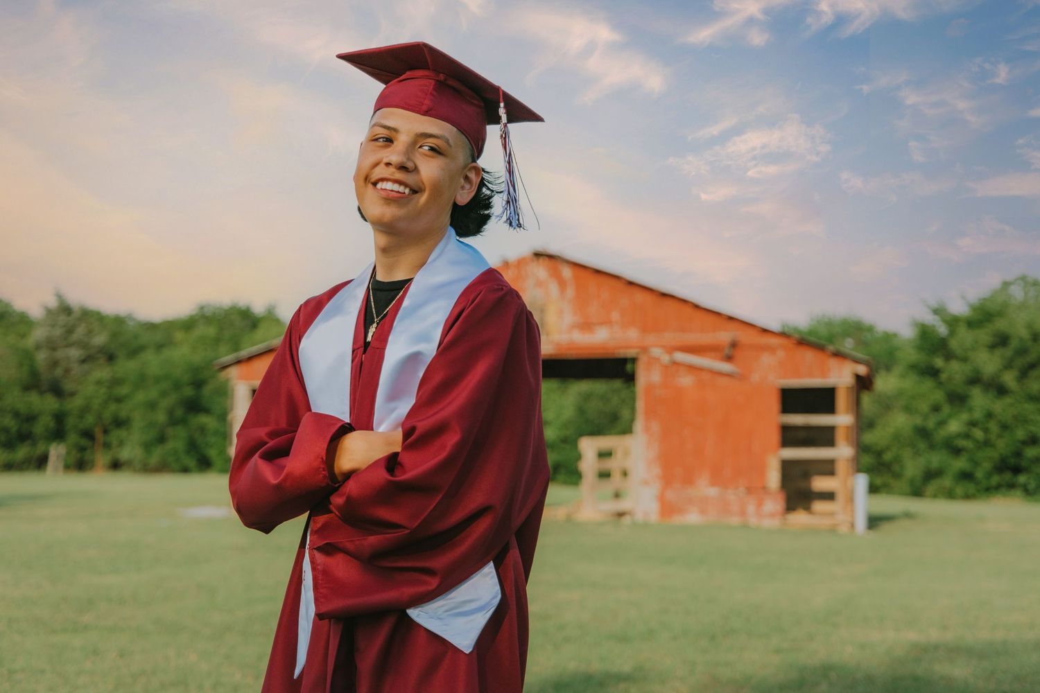 Graduate in red cap and gown stands arms crossed smiling in front of rustic barn outdoors.