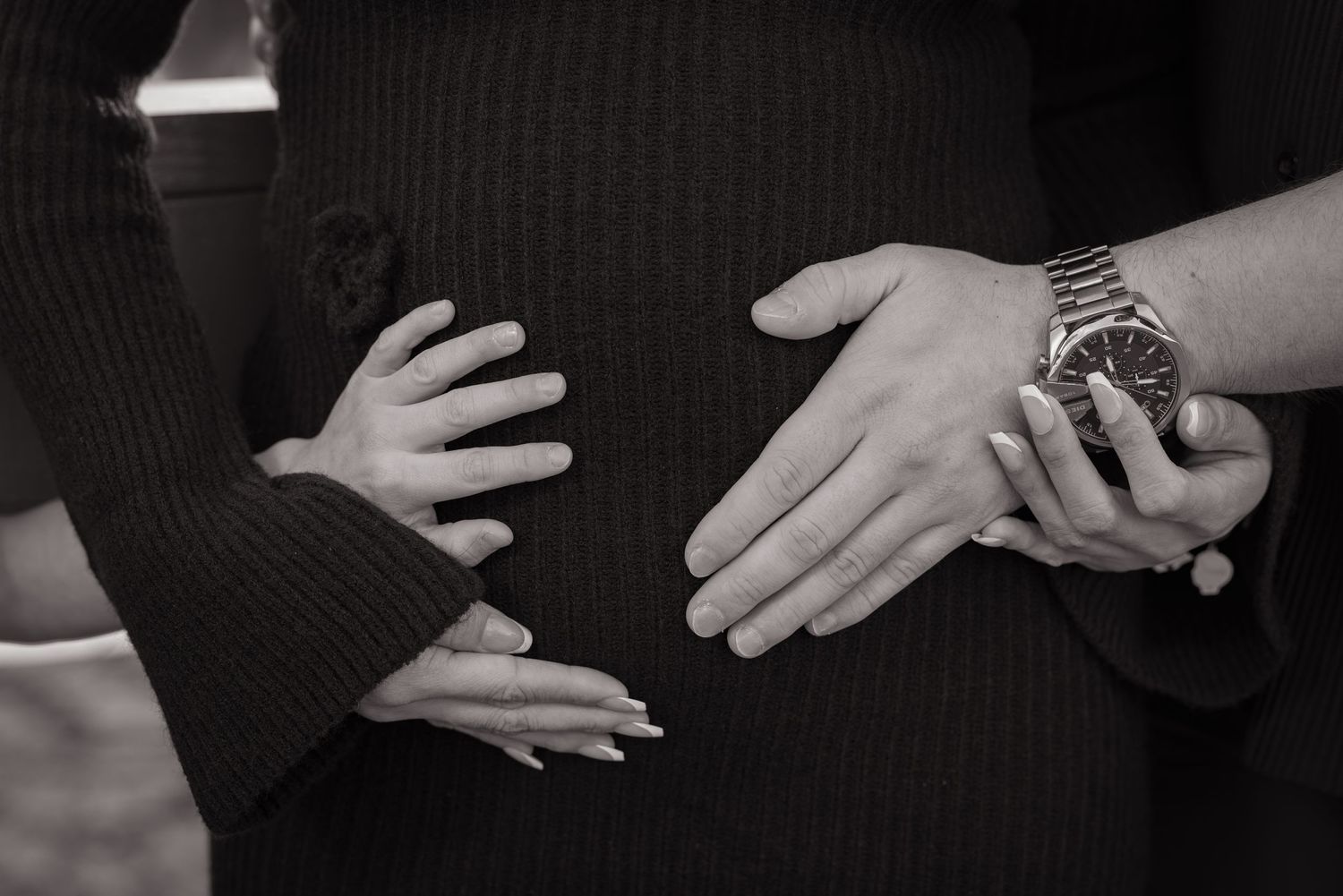 Black and white photo of hands on pregnant belly, including a child's hand, adult woman's hand, and man's hand with watch.