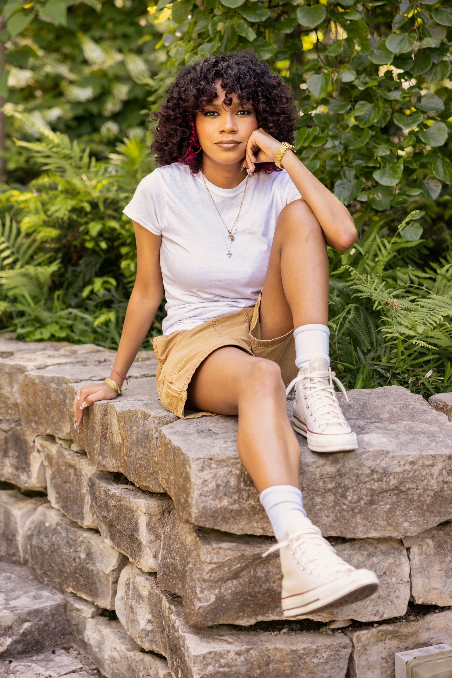 Young woman with curly hair sitting on stone wall in casual outfit for senior photos.