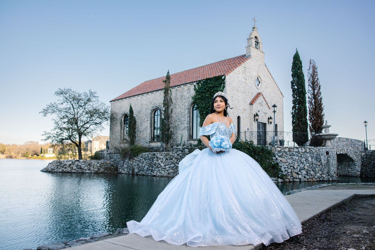 Young woman in blue quinceañera ball gown posing by a stone chapel beside a lake.
