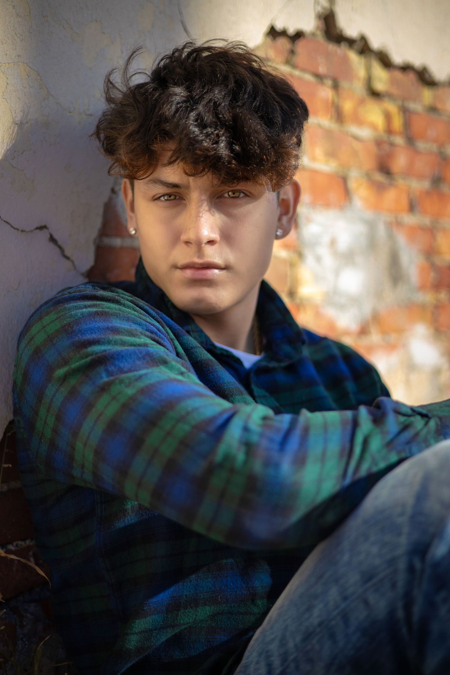 Young man in plaid shirt leans against cracked brick wall in moody portrait photo.