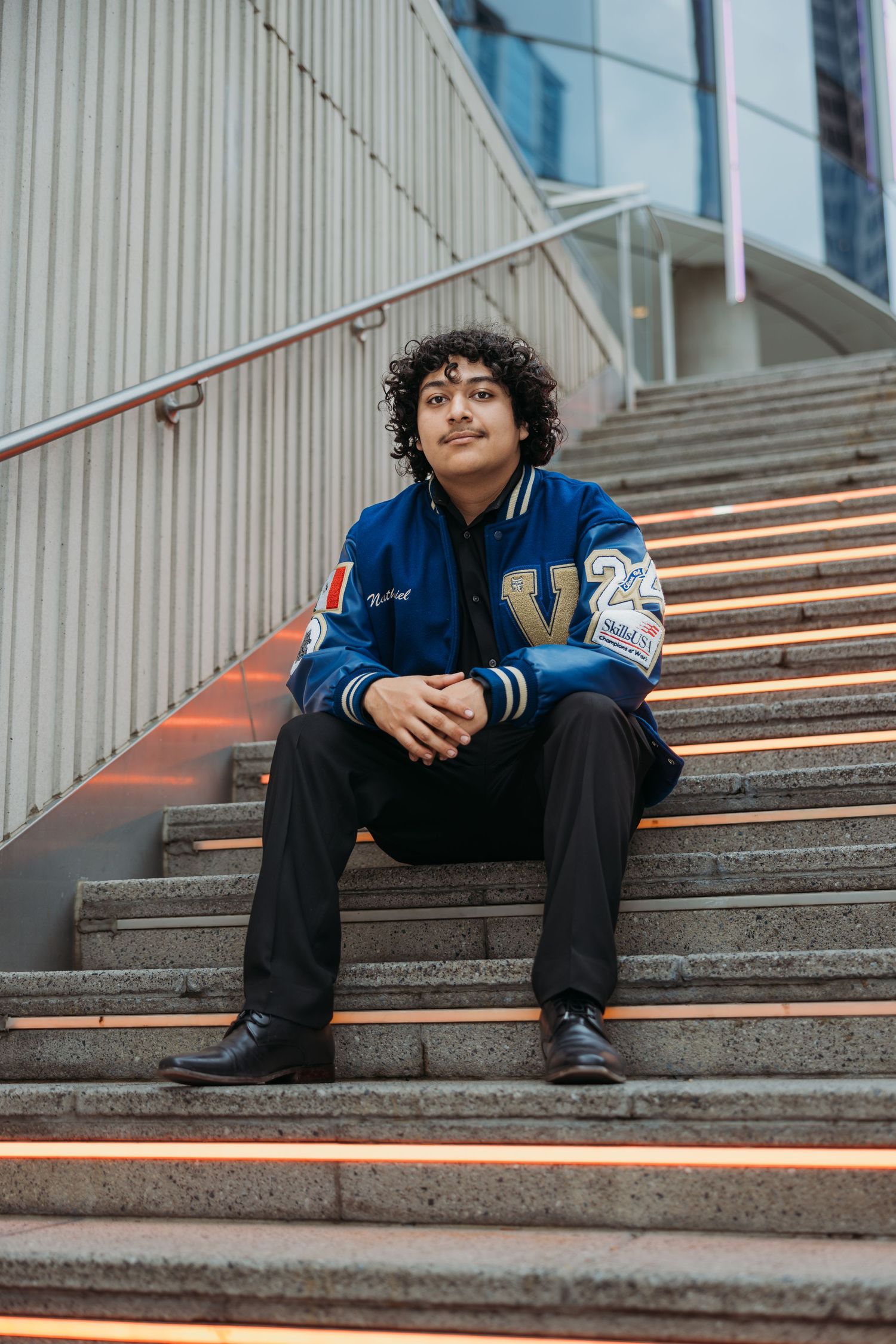 Young man in blue varsity jacket sitting on illuminated urban staircase steps.