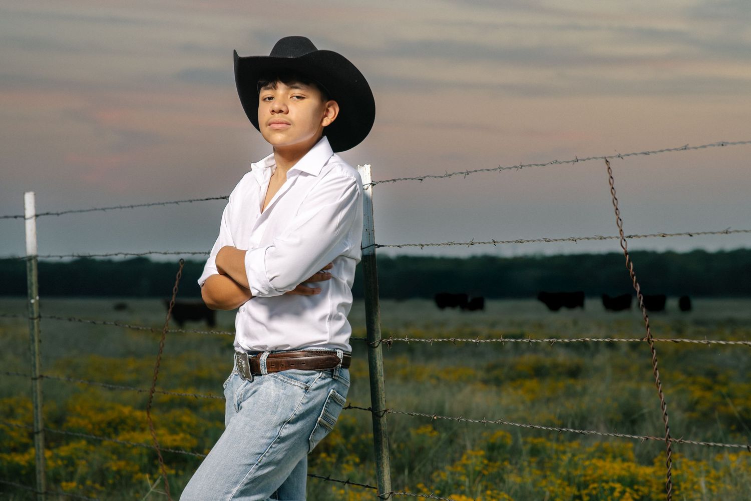 Young man in black cowboy hat and white shirt leaning on barbed wire fence with cattle grazing at sunset.