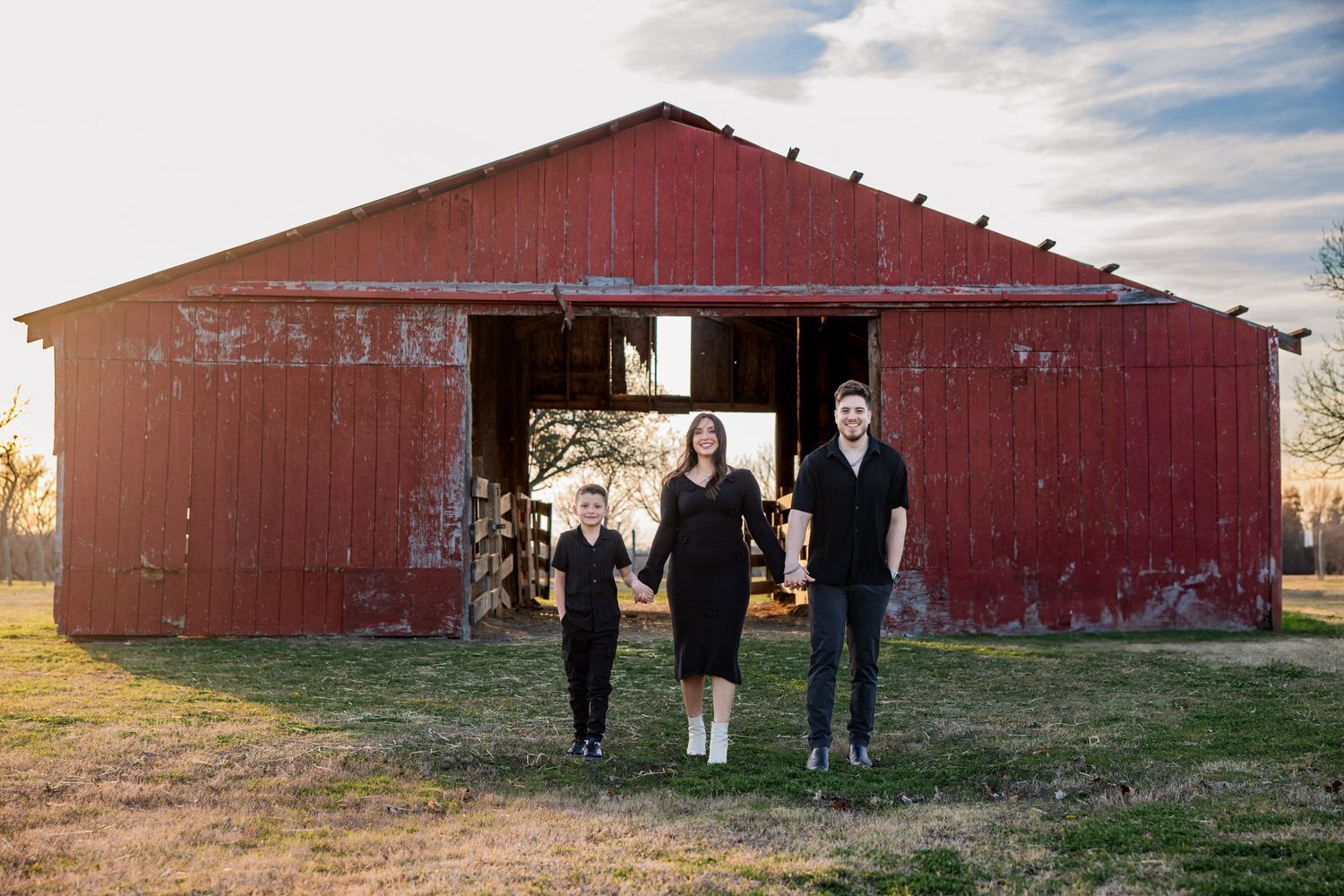 Family of three walking hand in hand in front of a rustic red barn at golden hour.