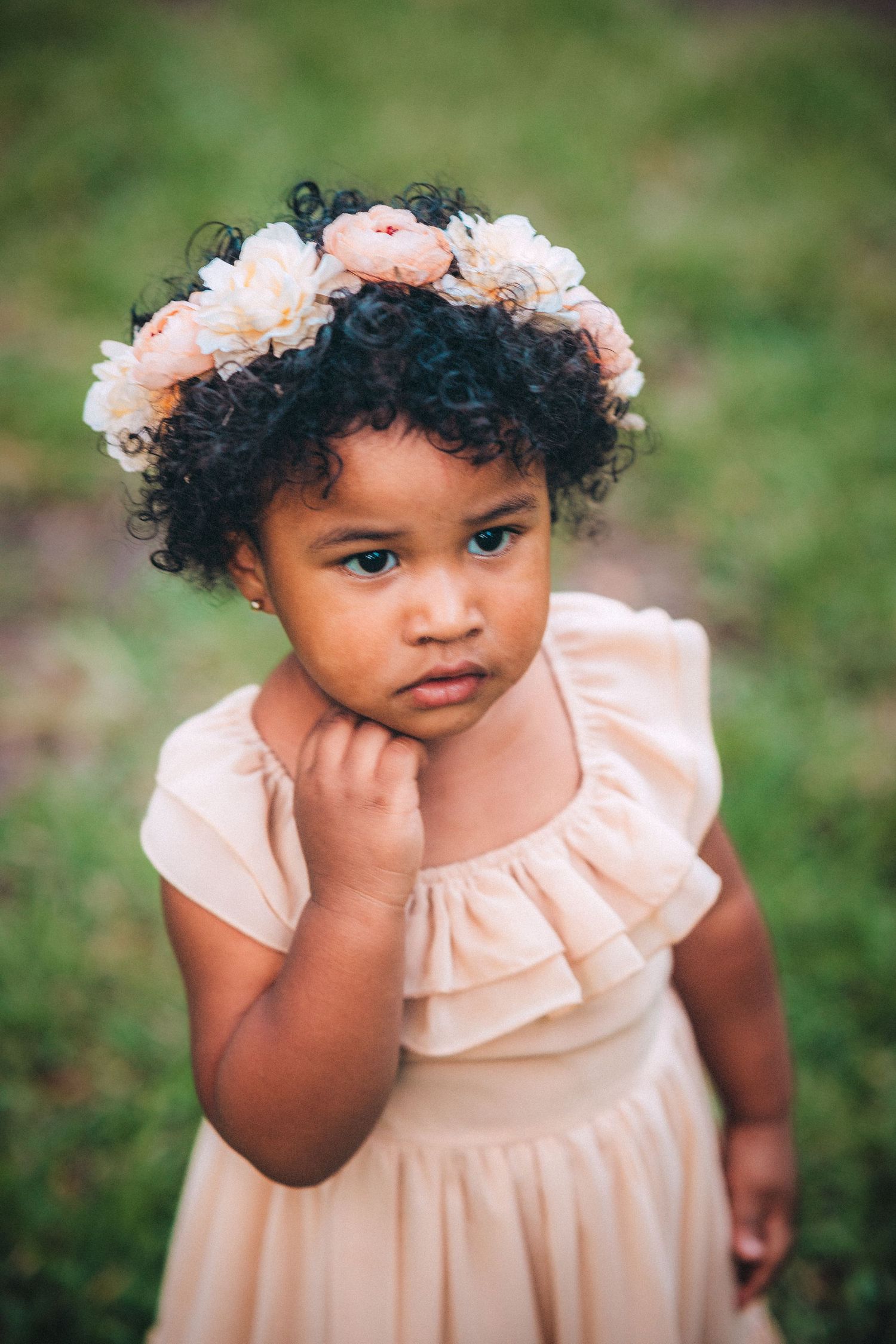 An adorable toddler girl in a blush dress wearing a floral crown poses outdoors in a garden.