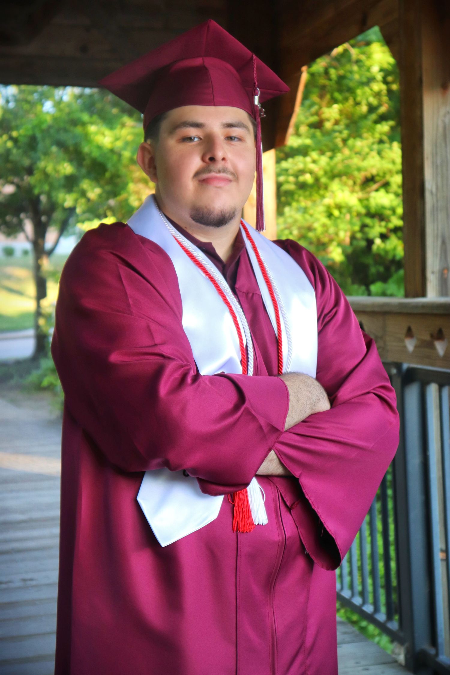Young male graduate in maroon cap and gown posing with arms crossed under a wooden pavilion.