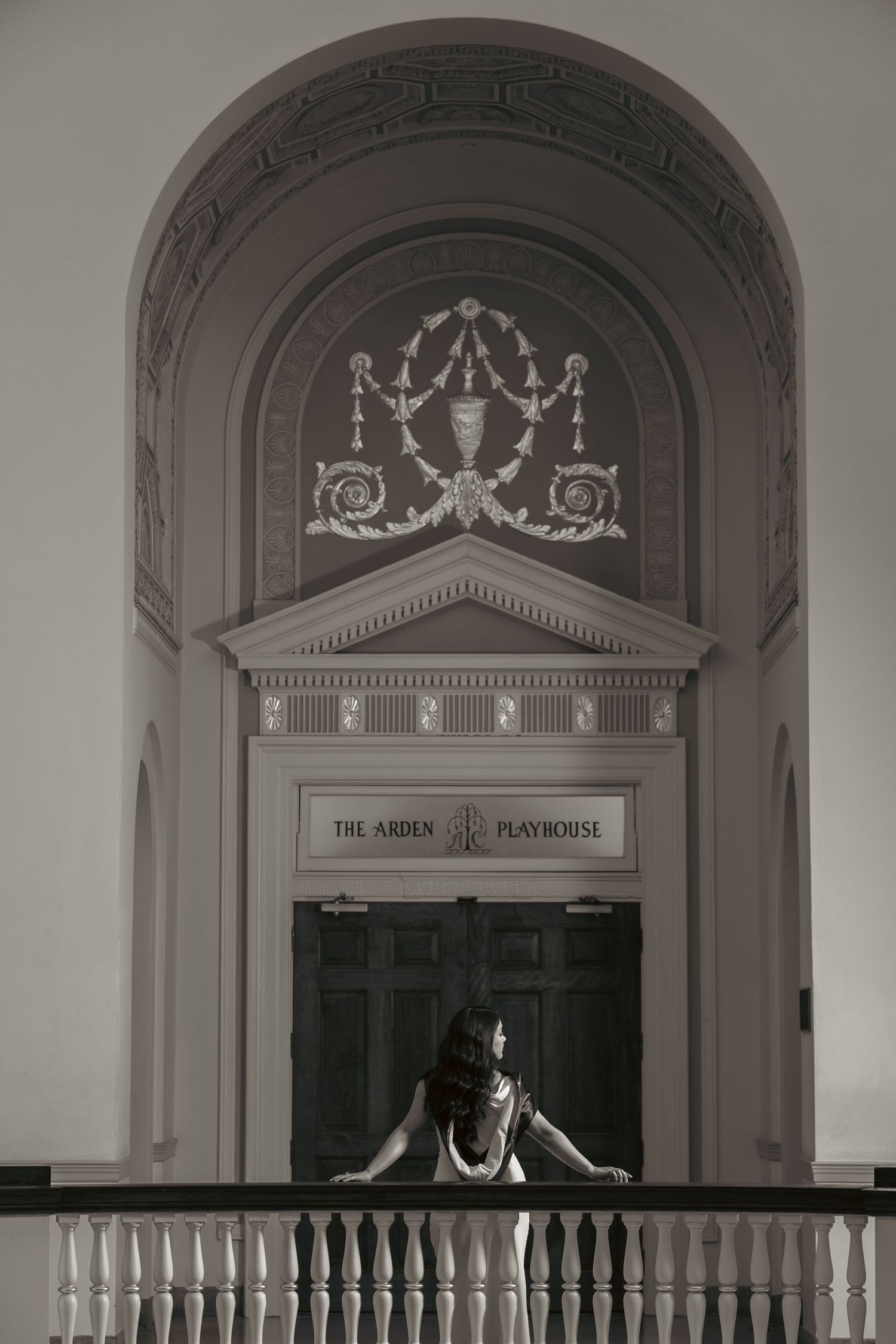 Woman posing at balcony railing before ornate arch entrance of The Arden Playhouse in black and white.